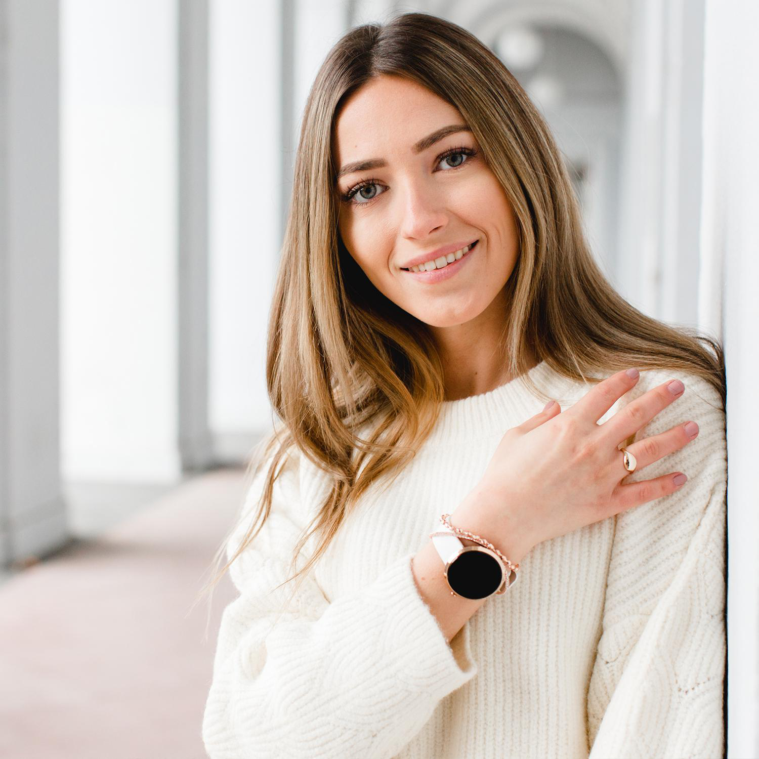 Lächelnde Frau, trägt einen weißen Pullover, lehnt an einer weißen Wand. Smartwatch und Armband sind sichtbar.