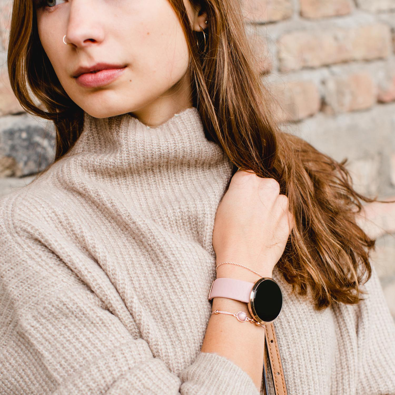 Eine Frau mit beigem Pullover und pinker Smartwatch mit passendem Armband, vor einer Backsteinmauer.