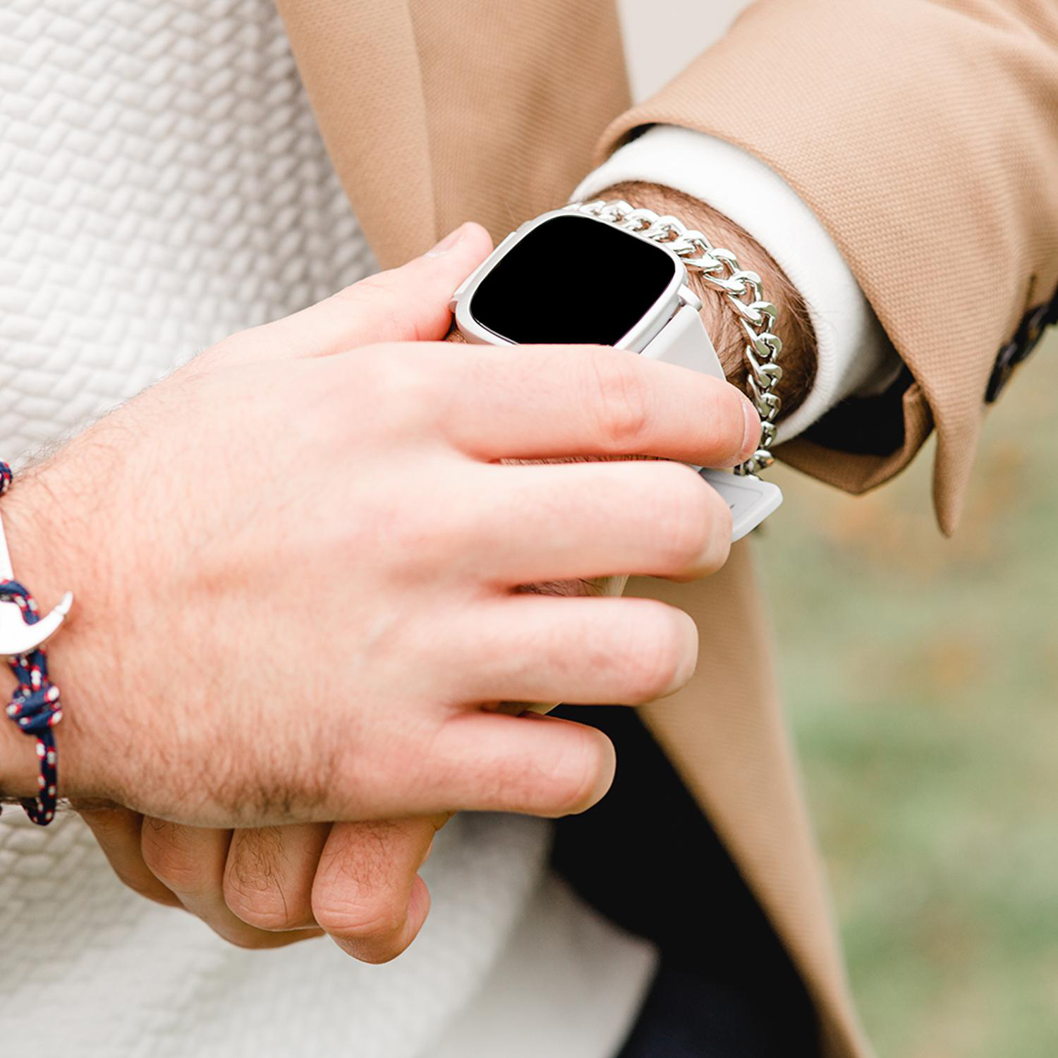 Eine Person im Anzug überprüft eine silberne Smartwatch und ein Kettenarmband. Der Hintergrund ist unscharf.