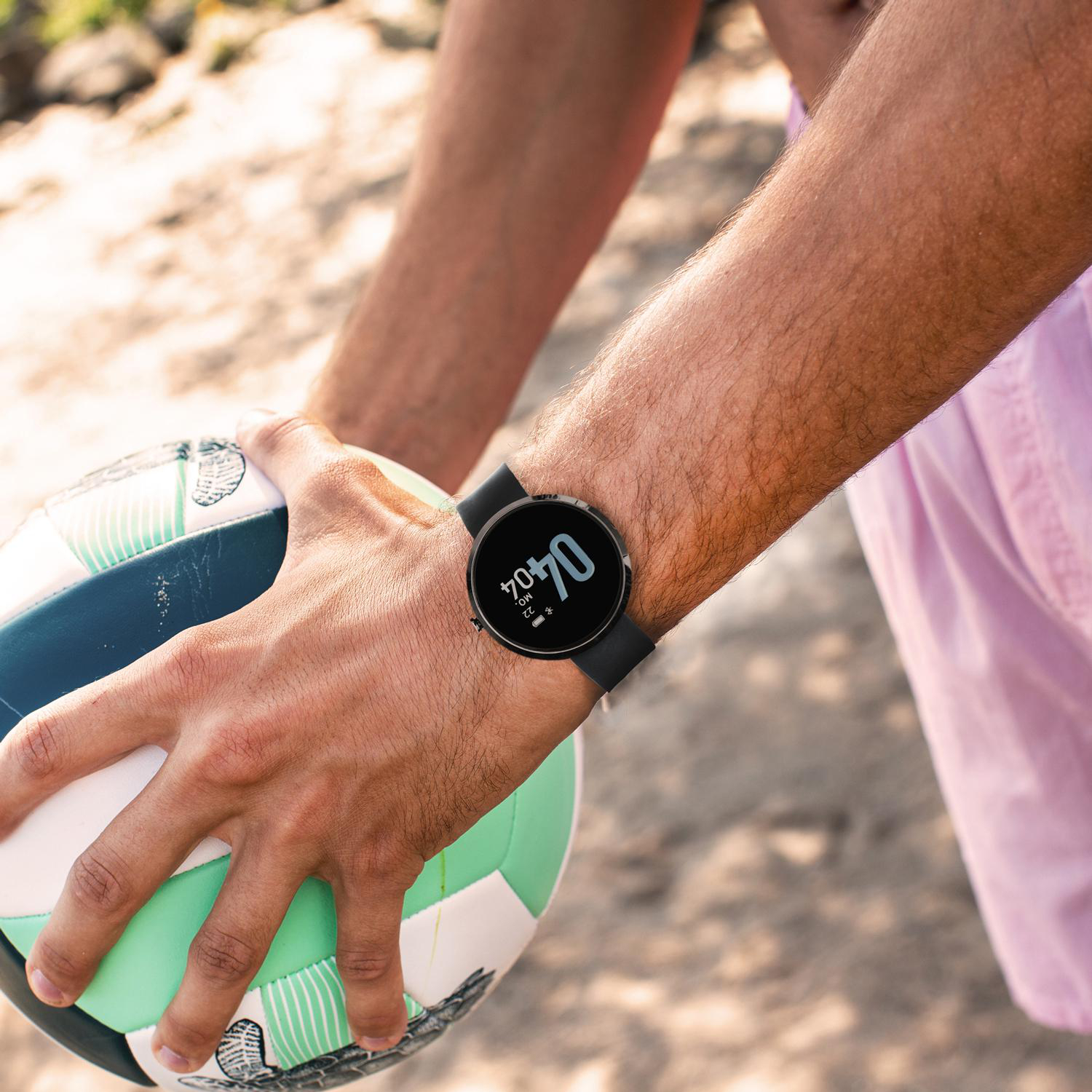 Die Hand einer Person, die eine Smartwatch trägt. Ein grün-blauer Ball auf dem Sand.