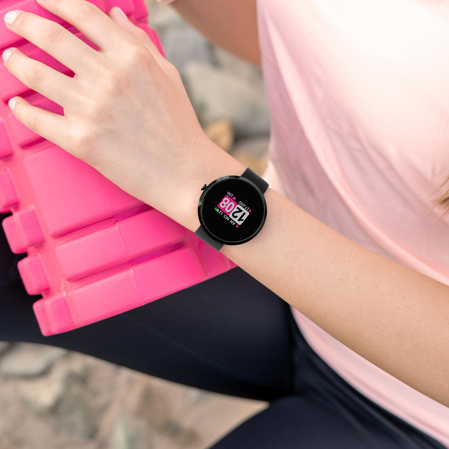 Das Handgelenk einer Frau mit einer schwarzen Smartwatch. Die Uhr zeigt Zeit und Datum an. Sie trägt ein rosa Hemd.
