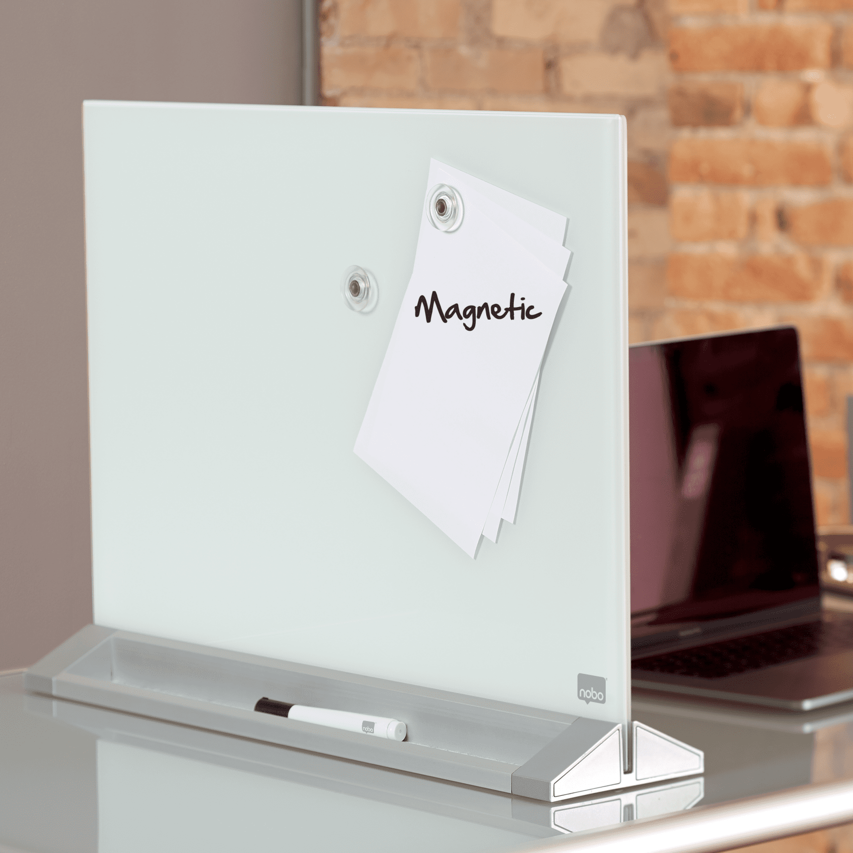 Üvegtábla papírokkal, tollal és laptop a háttérben. Téglából készült fal a háttérben.
