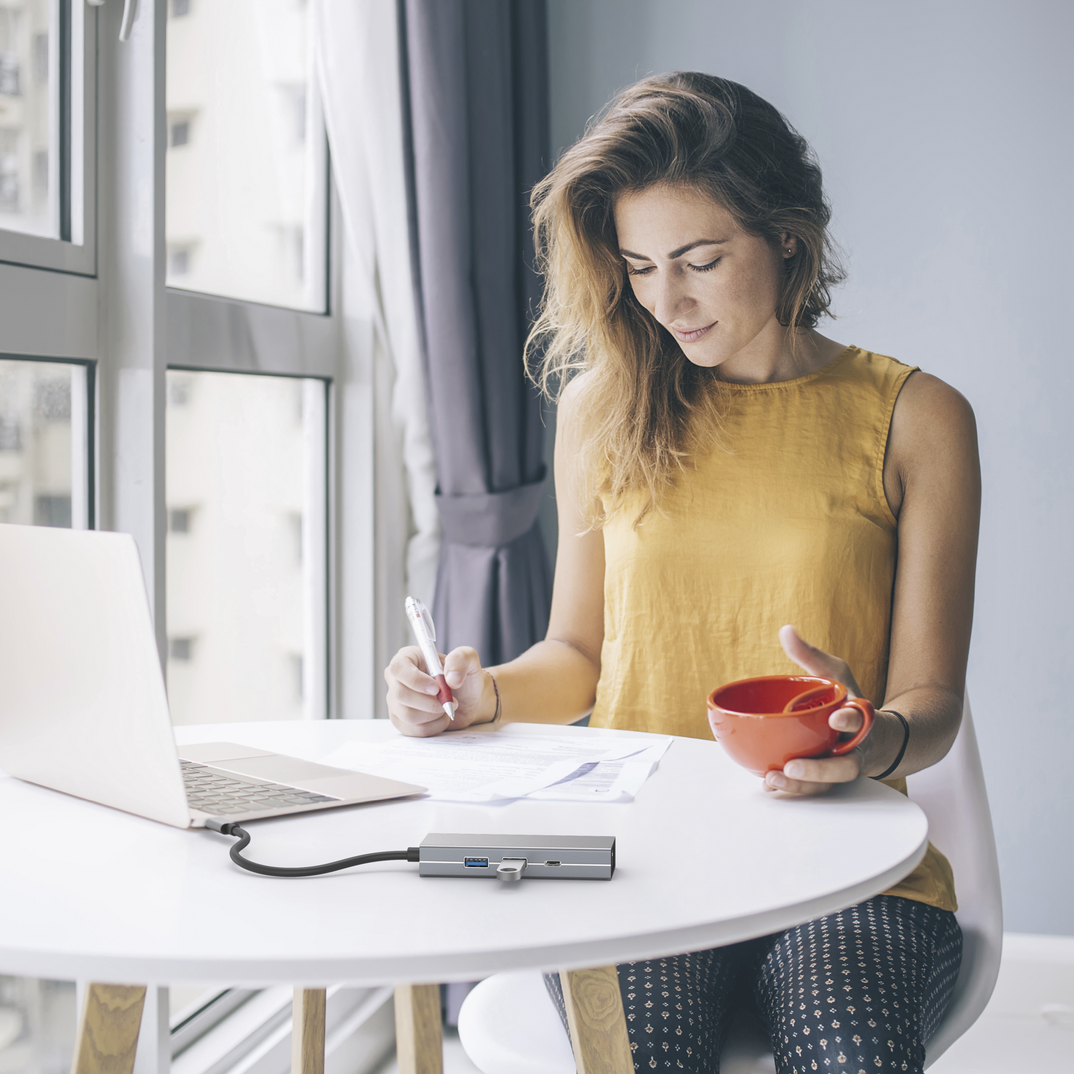 Mujer trabajando con portátil, café y hub en una mesa. Espacio de trabajo moderno, colores claros.