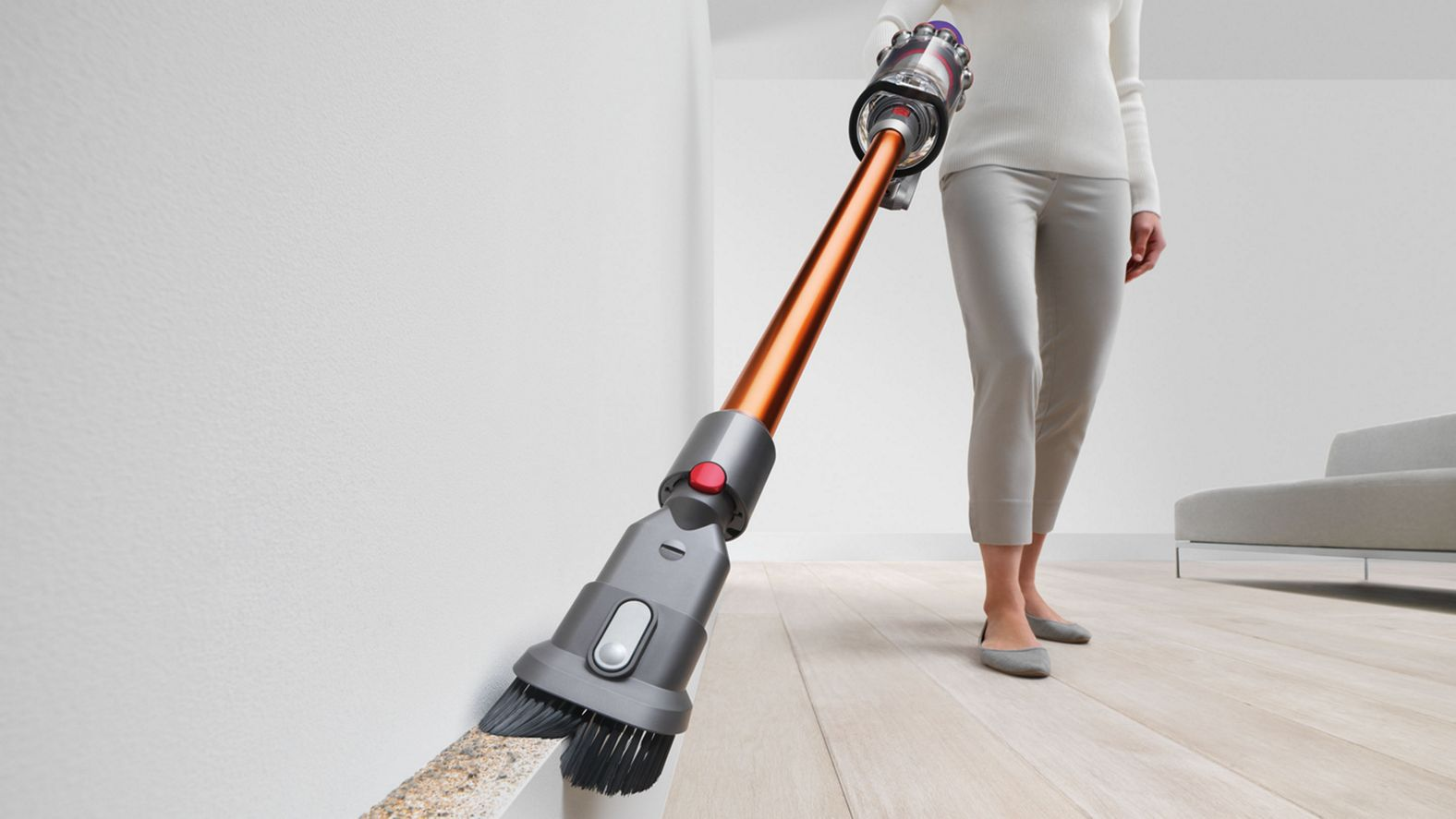 Eine Person saugt eine Wandecke mit einem orangefarbenen Staubsauger auf einem Holzboden.