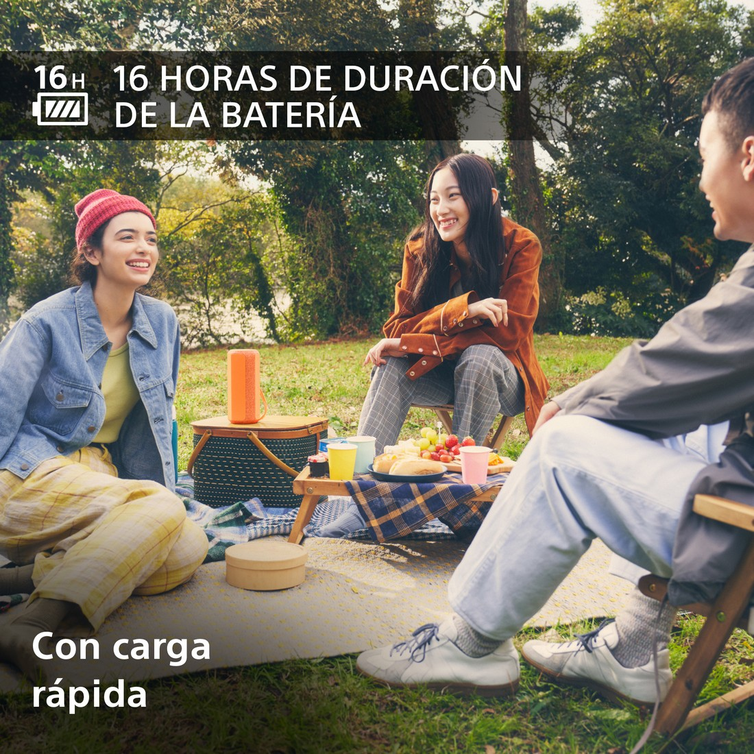 Tres personas sentadas en una manta en un parque. Hay comida y un altavoz en la manta.