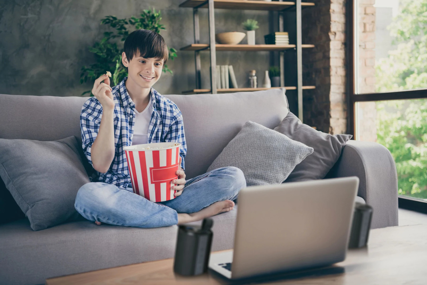 Ein junger Mann sitzt auf einem Sofa, schaut auf einen Laptop und isst Popcorn.