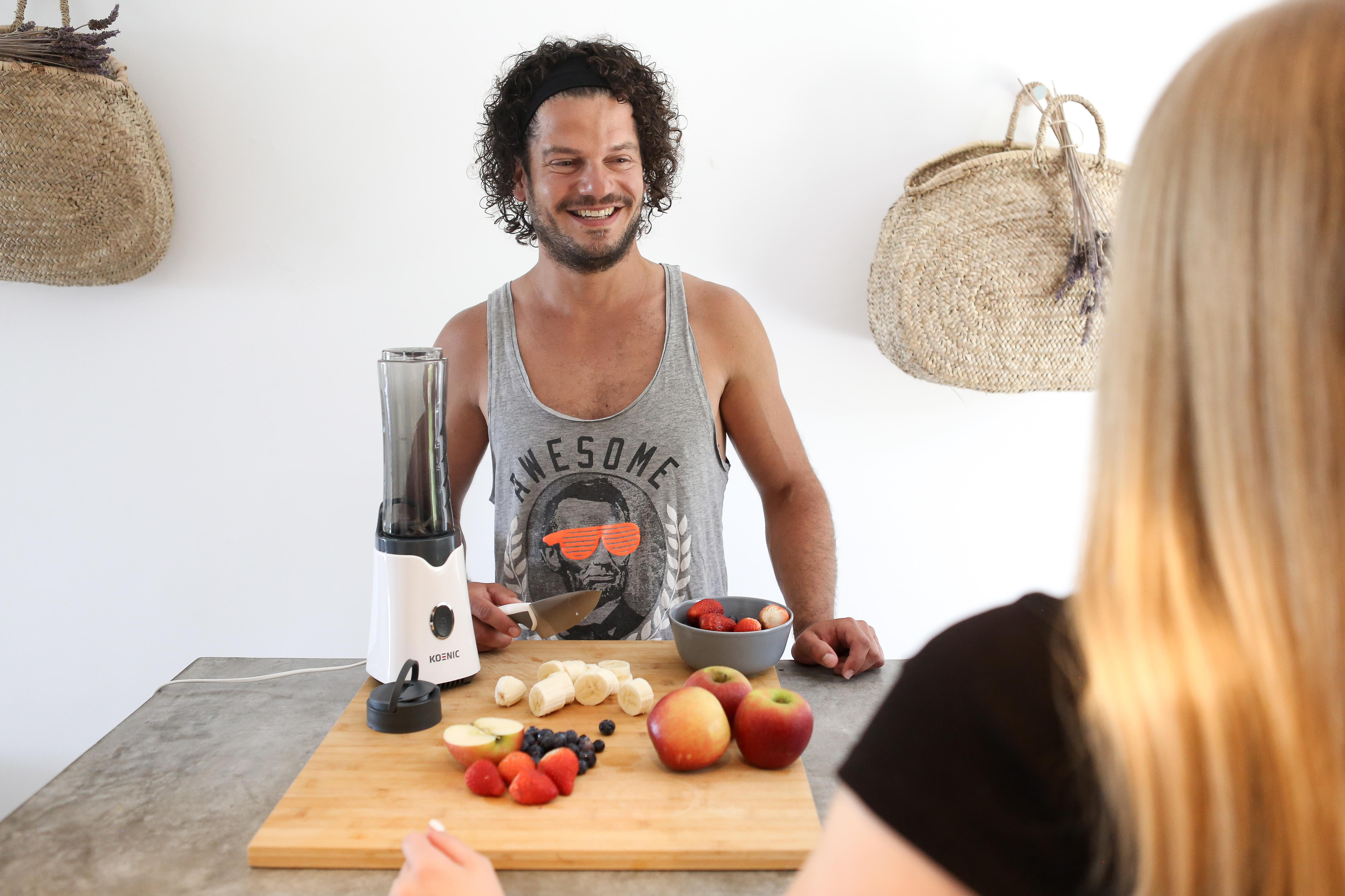 Mann lächelt, steht hinter einer Theke mit Früchten und Mixer. Eine andere Person im Vordergrund.