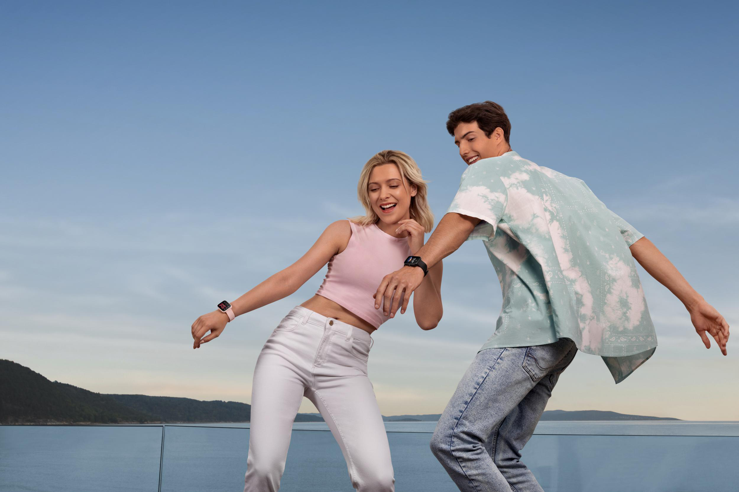 Zwei tanzende Personen mit Smartwatches, mit sonnigem Himmel und Wasser im Hintergrund.