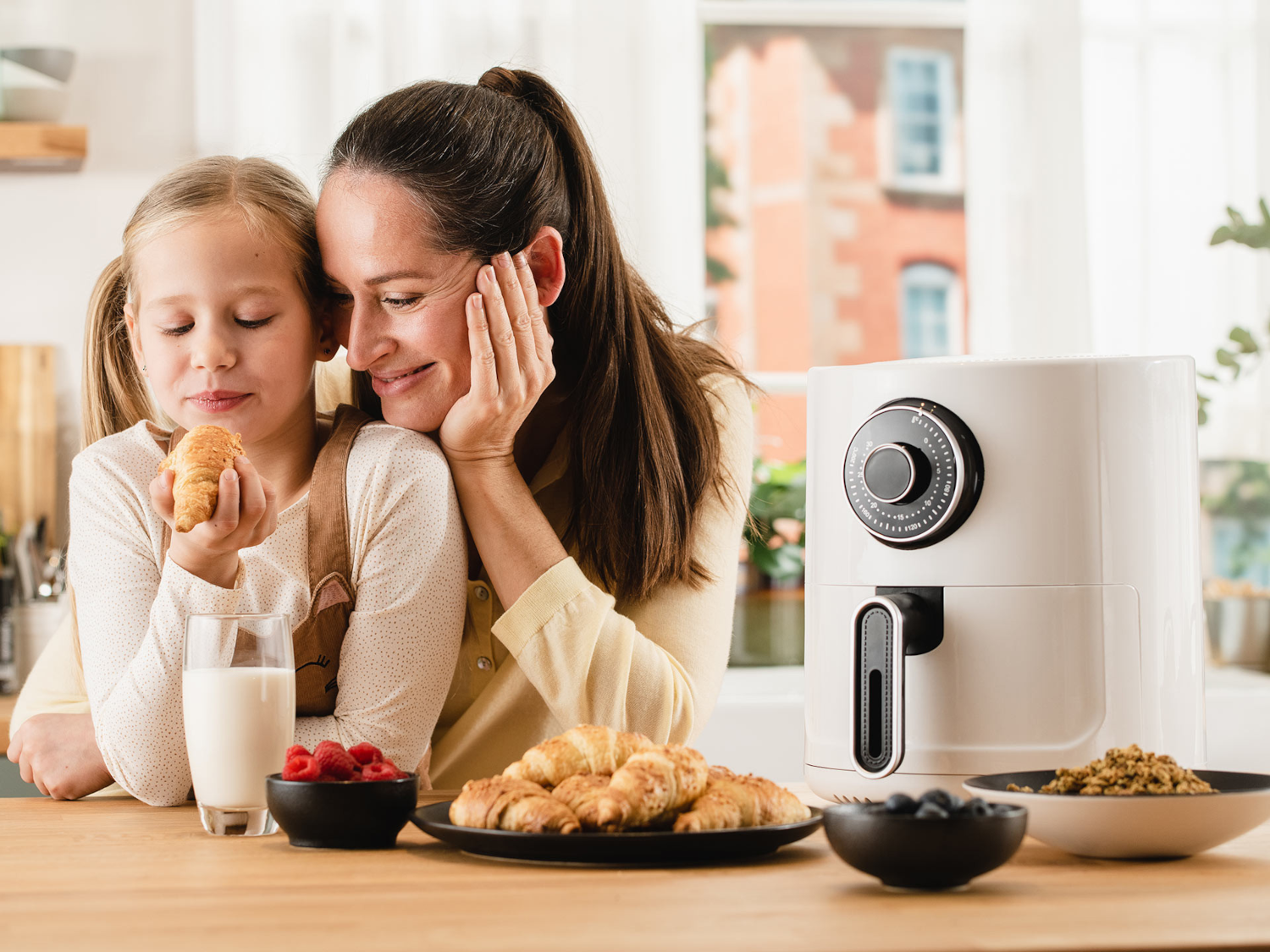 Egy anya és lánya a konyhában. A lánya egy croissant-t tart. Egy fehér air fryer van a közelben.