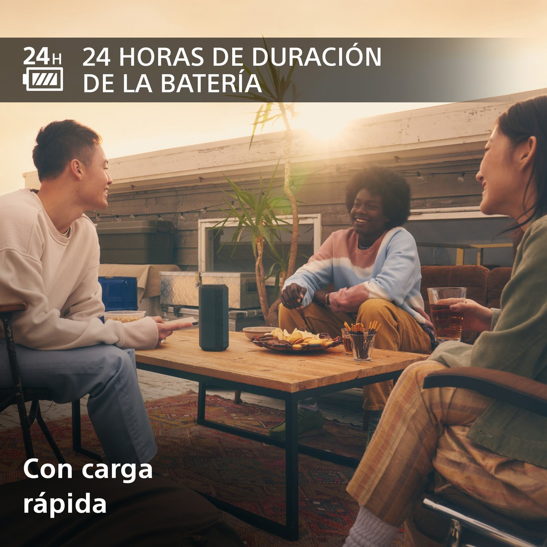 Tres personas en una azotea, socializando. Hay un altavoz sobre la mesa y el texto dice batería de 24 horas.