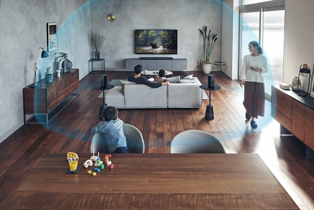 Una escena de sala de estar con gente viendo la televisión, con altavoces. Una mesa de comedor con juguetes está al frente.