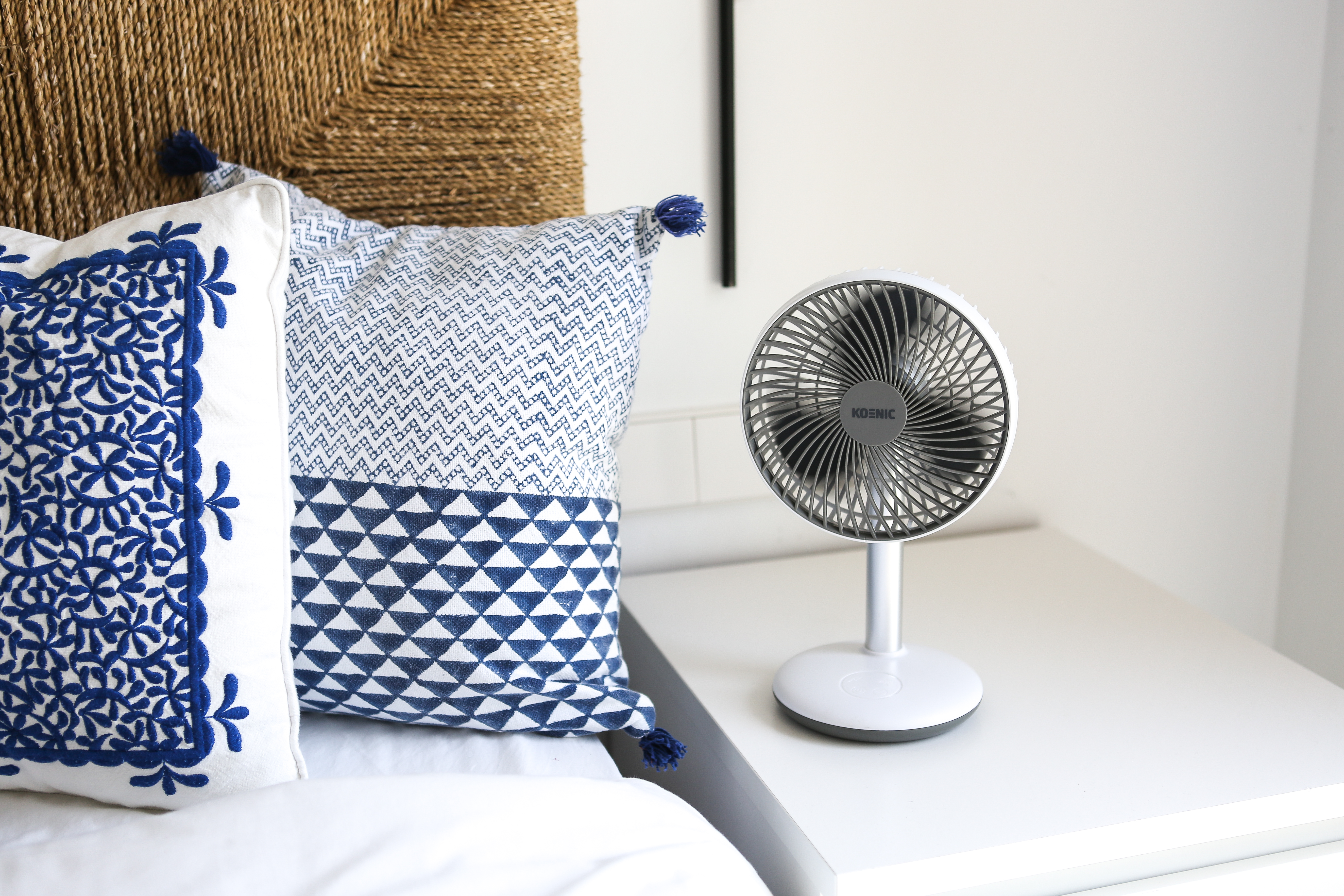 Weißer Ventilator auf weißem Tisch, blau-weiße Kissen und ein Hintergrund mit Flechtmuster.