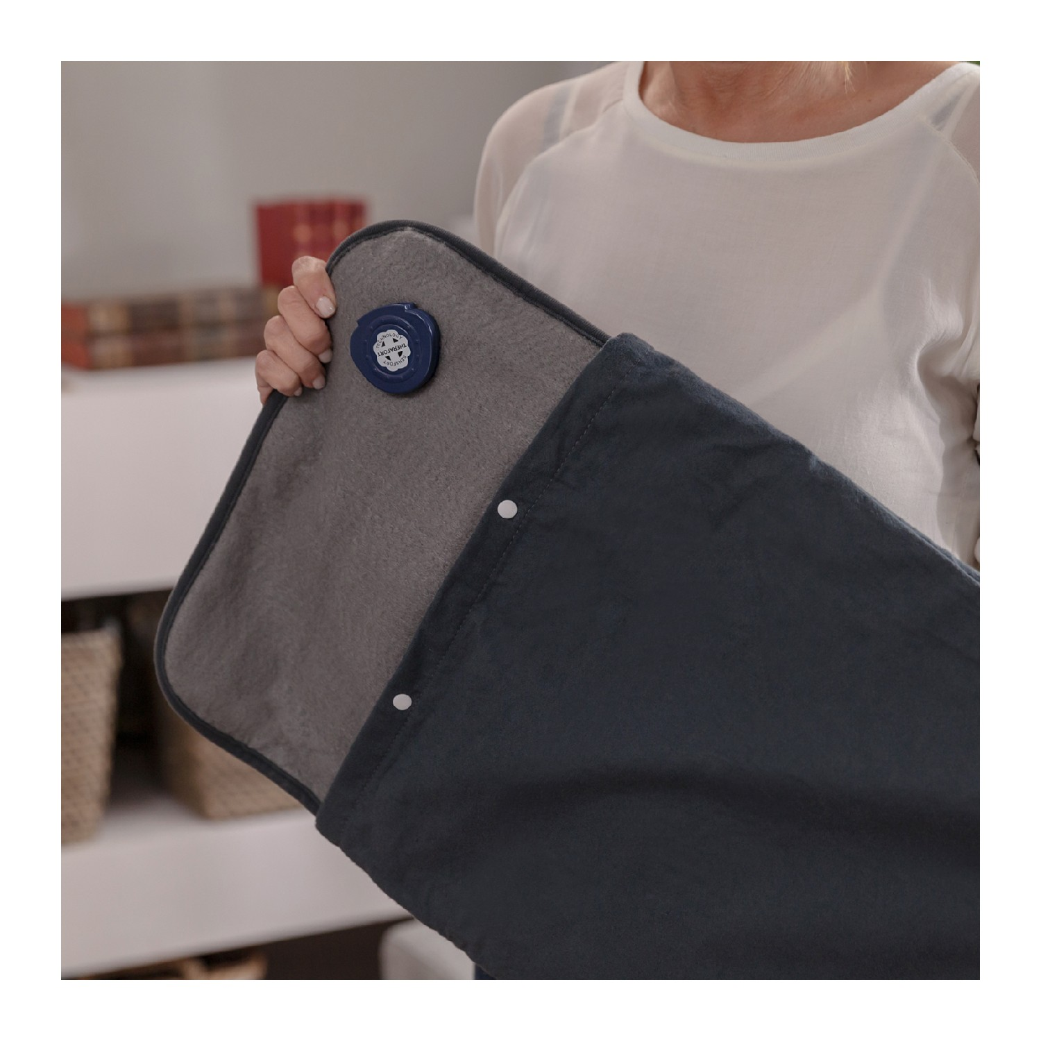 Una persona sostiene una manta térmica azul oscuro con una capa interior gris. Un botón de control azul es visible. El fondo está borroso.
