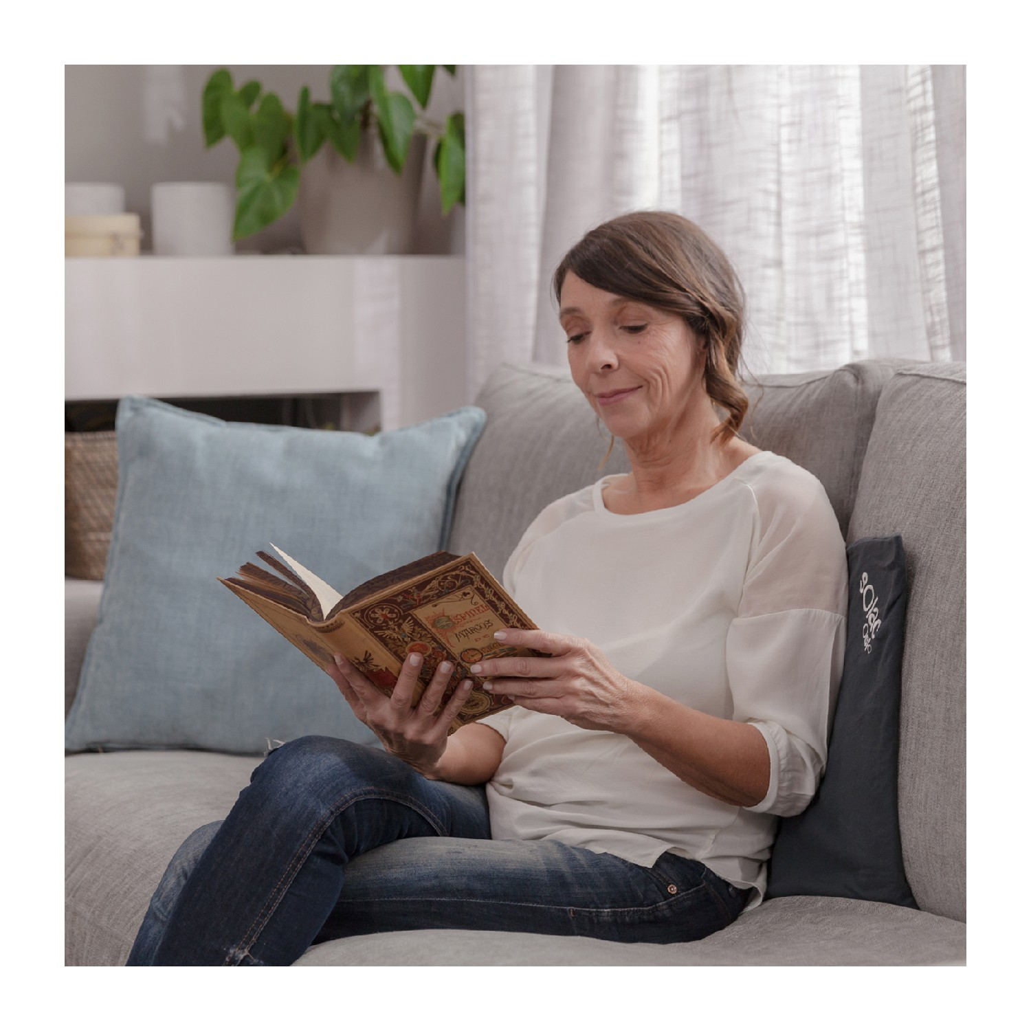 Una mujer sentada en un sofá lee un libro. Viste camisa blanca y vaqueros. Un cojín azul y plantas en el fondo.
