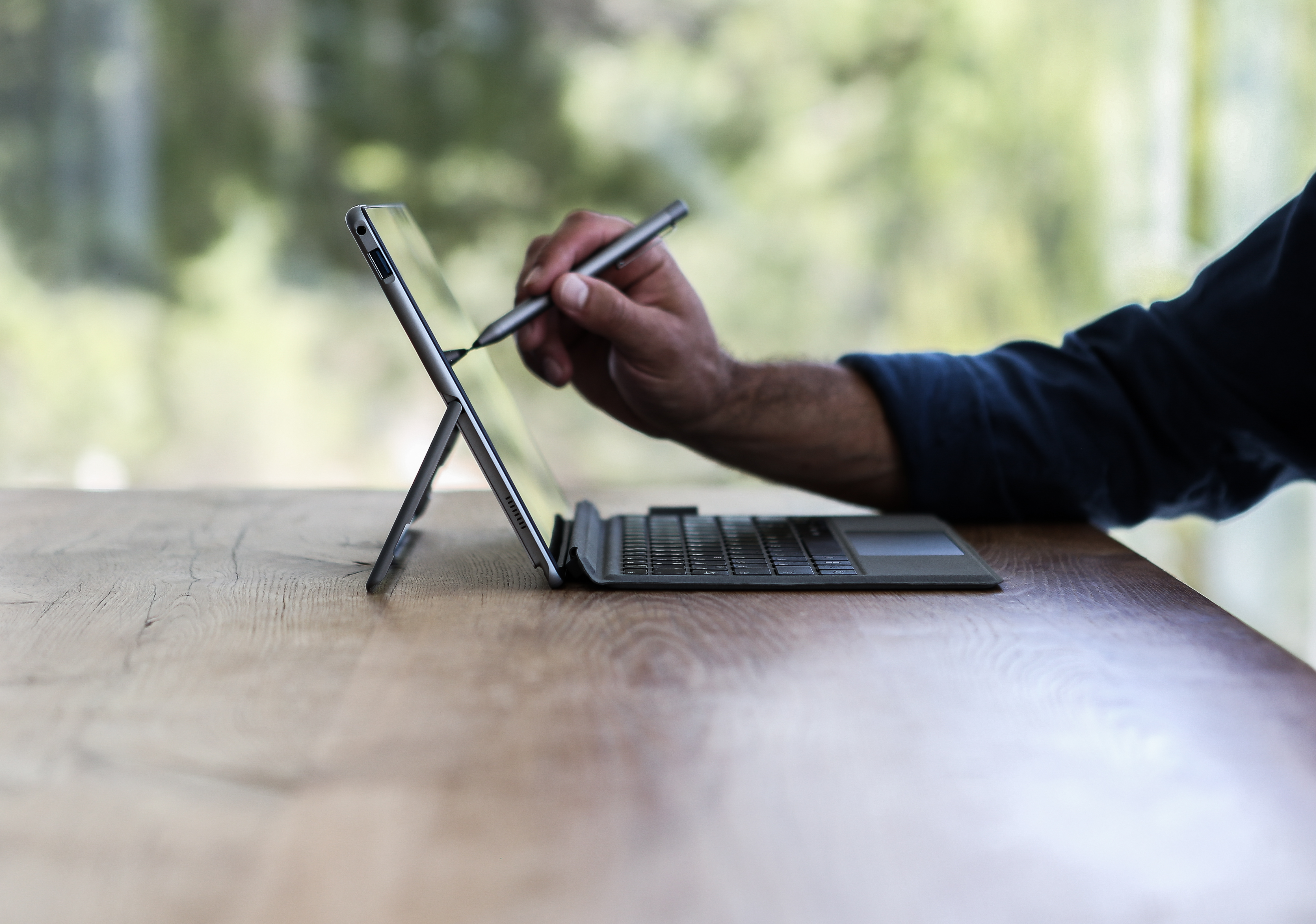 Person verwendet Stift auf Tablet mit Tastatur, auf einem Holztisch.