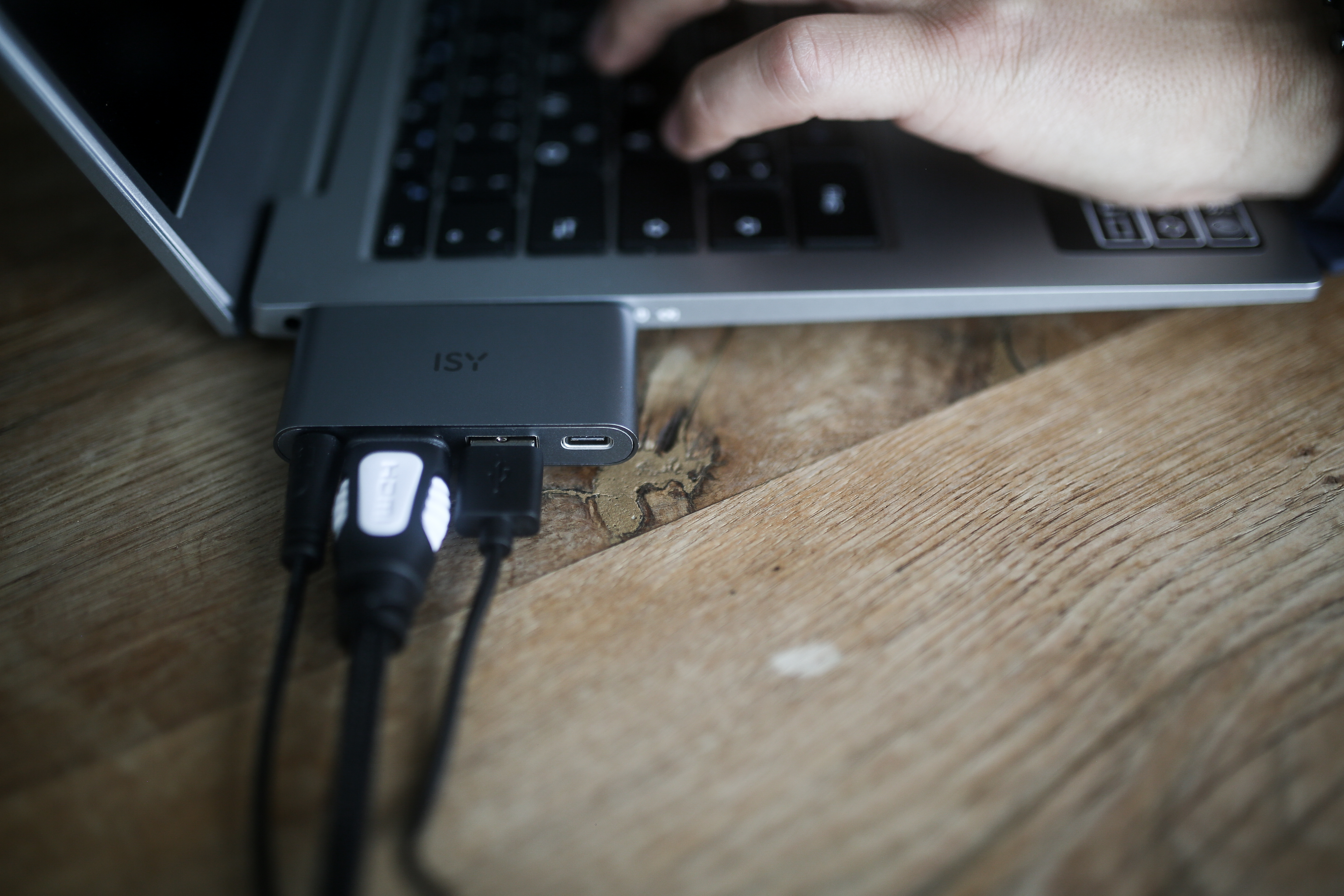 Laptop mit Hub und Kabeln auf Holztisch, Hand tippt.
