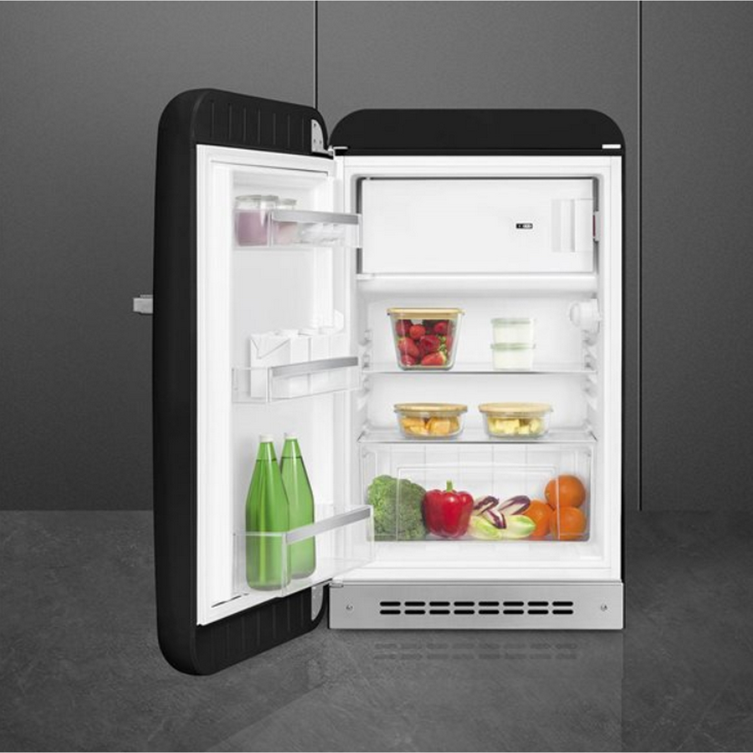 Refrigerador retro en blanco y negro, con la puerta abierta mostrando comida. Está en una habitación gris.