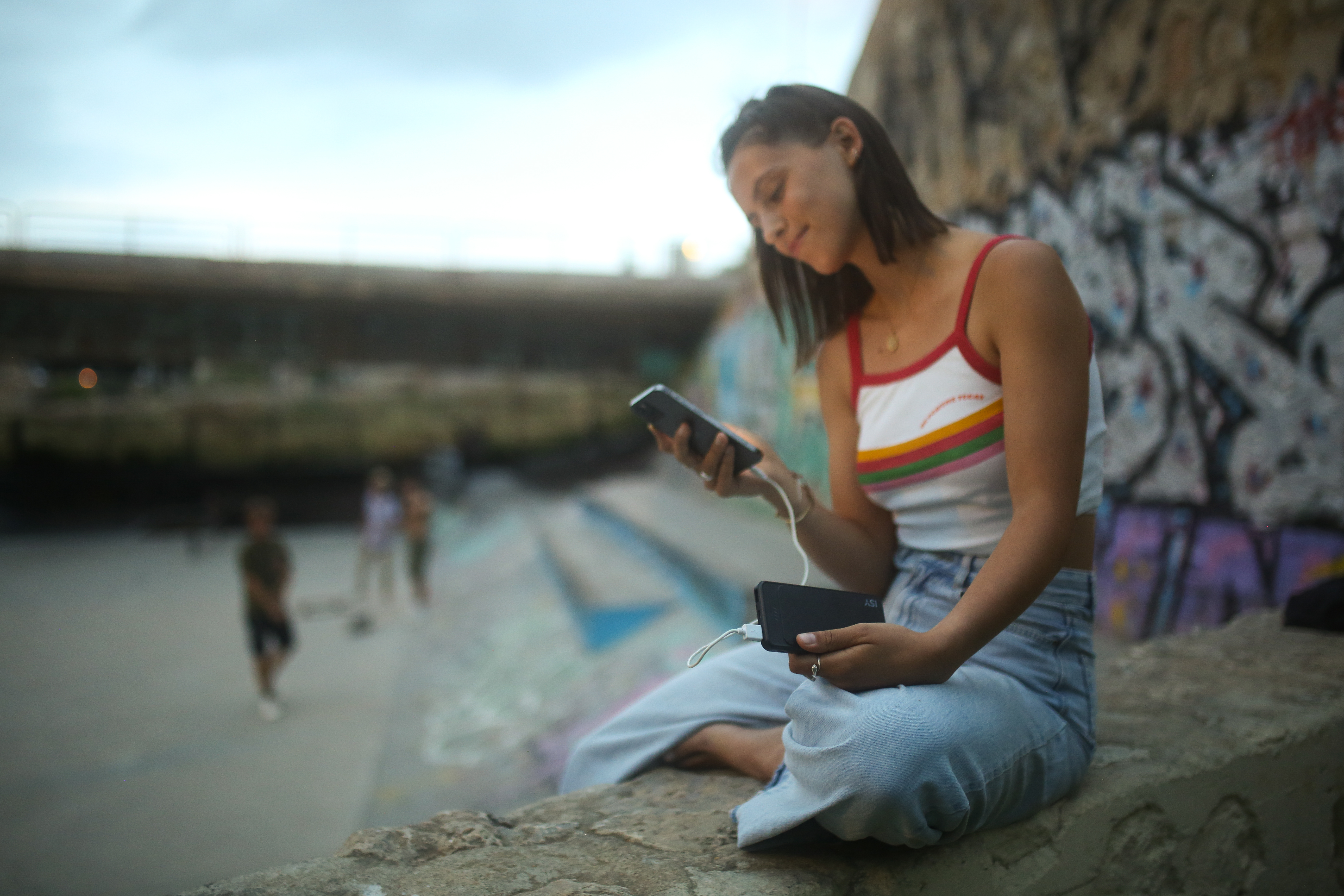 Frau lädt Telefon mit Powerbank auf, sitzt in der Nähe von Graffiti.