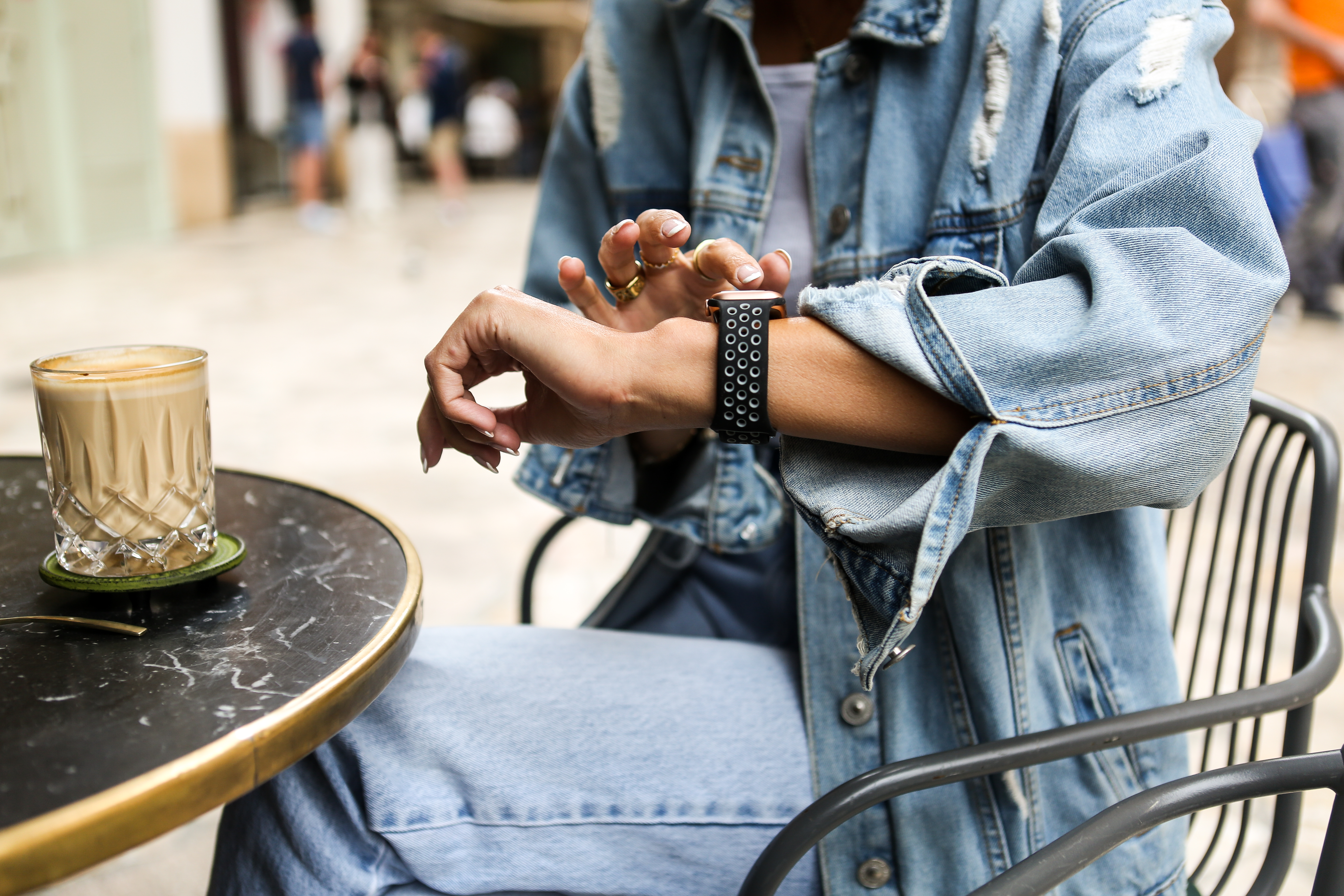 Person in Jeansjacke betrachtet Smartwatch, Kaffee auf dem Tisch.