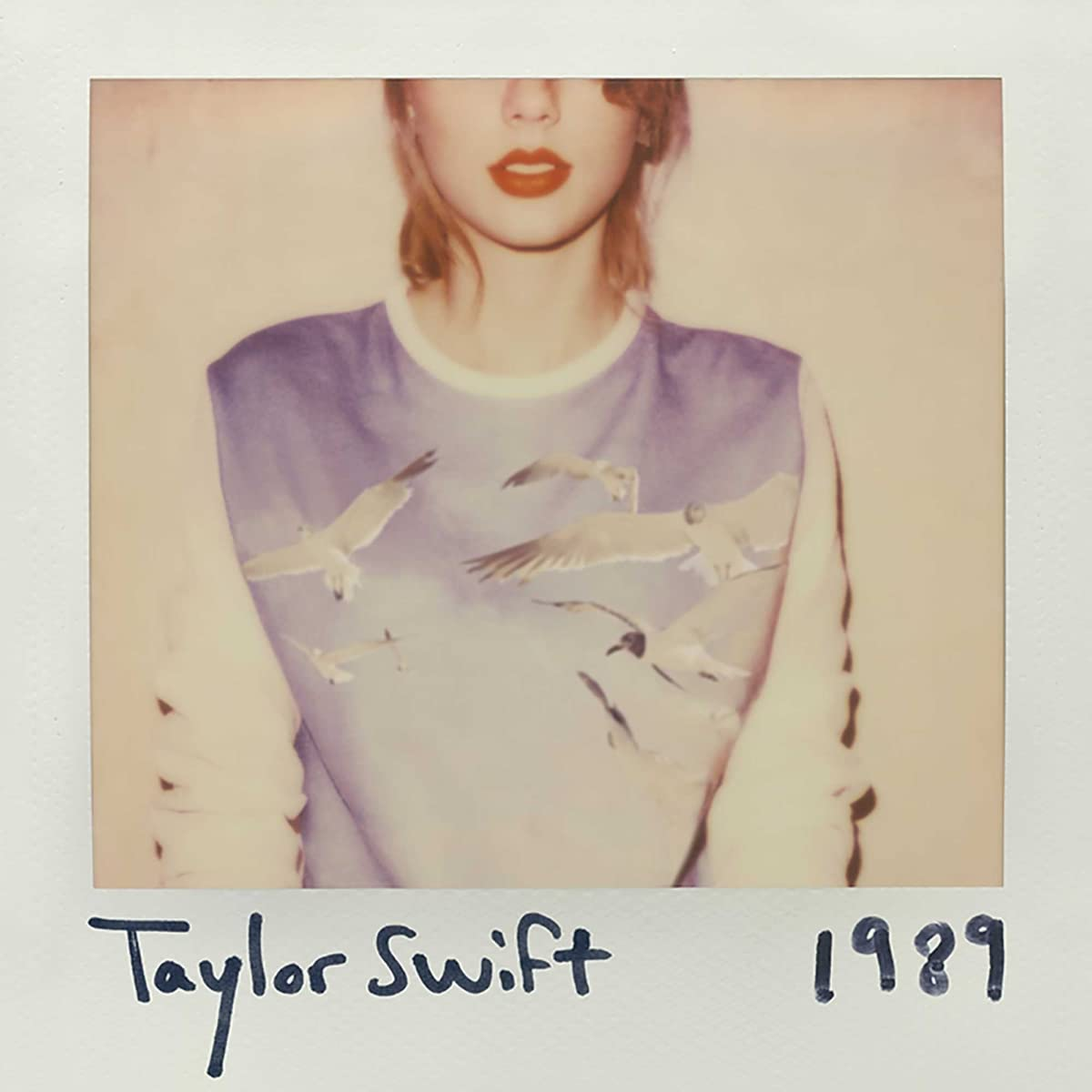 Polaroid fotó Taylor Swiftről, kék inget visel sirályokkal, fehér kerettel és fekete szöveggel.