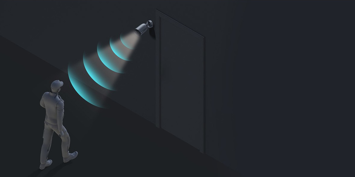 A security camera shines a beam of light on a man walking toward a door in a dark setting.