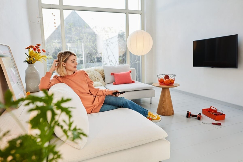 Nő a kanapén tévét néz, távirányítót tart. Asztal tálal és virággal mellette.