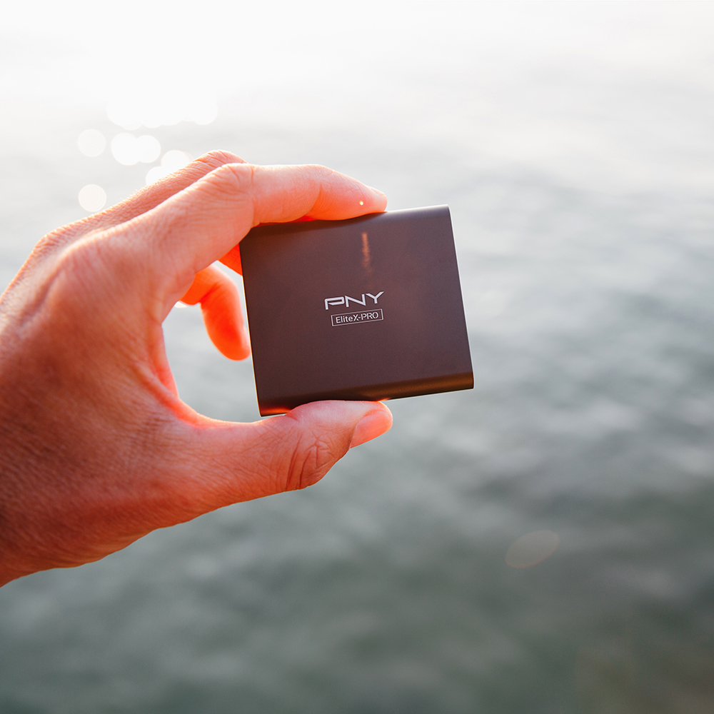 Hand holding a small brown square PNY Elite-PRO external hard drive, water in background.