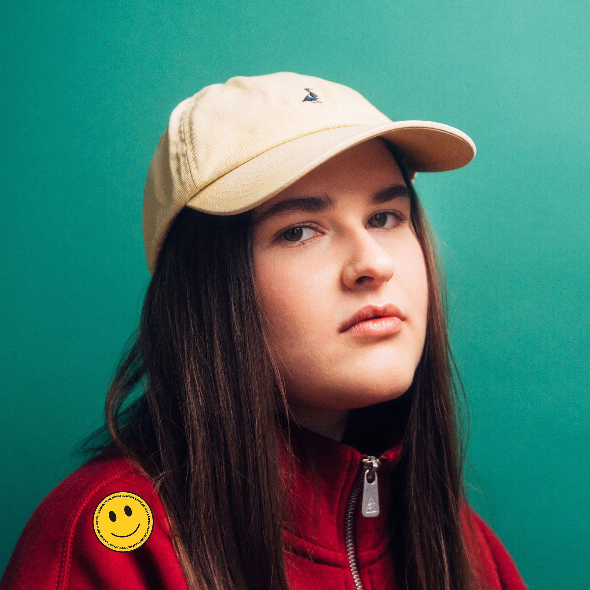 Eine junge Frau mit gelber Kappe und roter Jacke. Auf der Jacke befindet sich ein Smiley-Aufkleber.