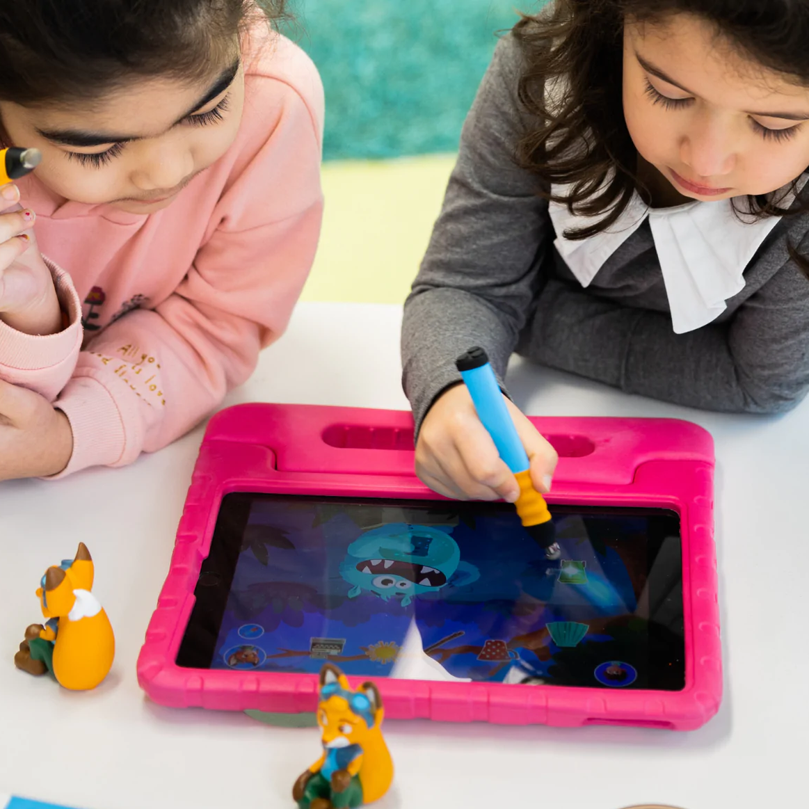 Zwei Mädchen zeichnen mit blauen Stiften auf einem rosa Tablet. Spielzeugfüchse sind vorhanden.