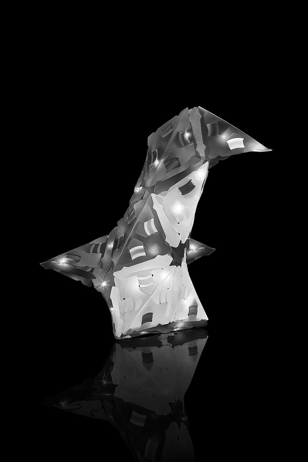 Graustufen, abstrakte Skulptur mit Lichtern, vor schwarzem Hintergrund, ähnelt einer stilisierten Form.