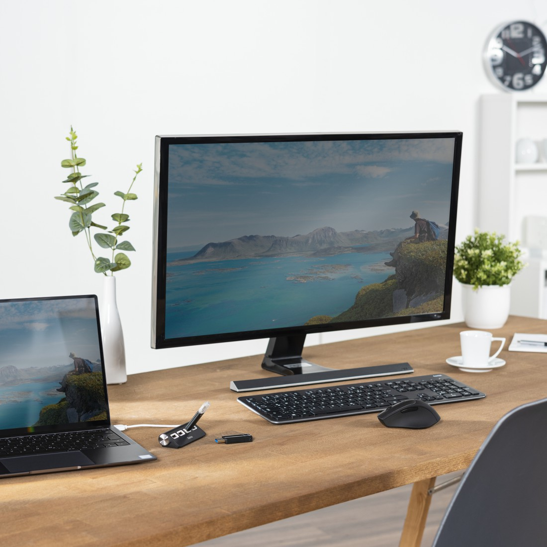 Escritorio con portátil, monitor, teclado, ratón y planta. Ventana al fondo.