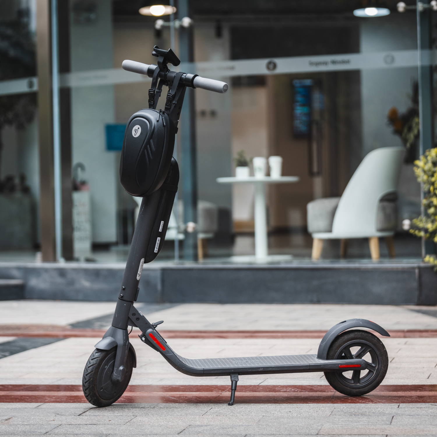 An electric scooter parked on a sidewalk. A black bag is attached to the handlebar, glass door background.
