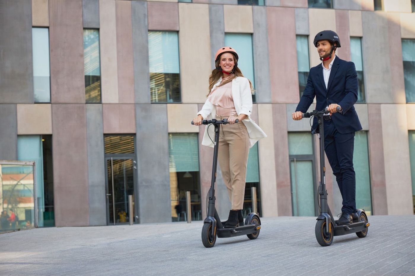 İki kişi, bir erkek ve bir kadın, asfalt bir alanda elektrikli scooter sürüyor. Kask takıyorlar.