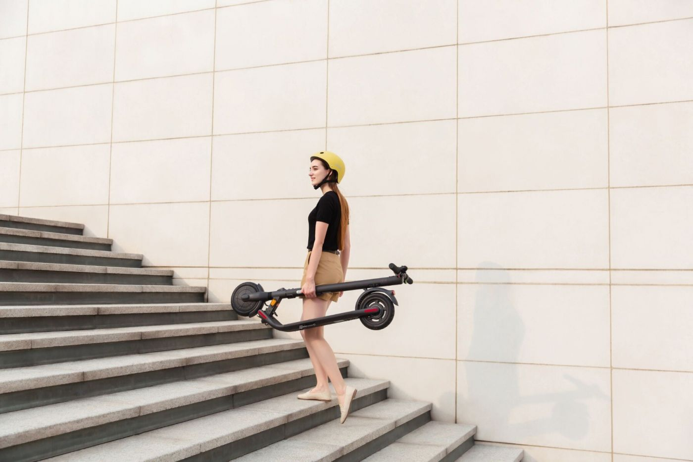 Bir kadın merdivenlerden katlanmış bir scooter taşıyor, kask takıyor.