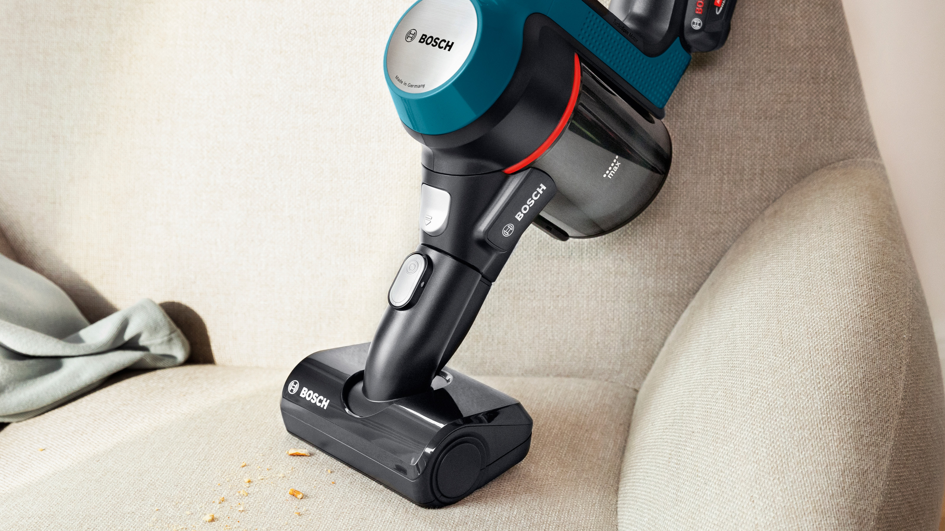 Ein blauer und türkisfarbener kabelloser Bosch-Staubsauger reinigt ein beiges Sofa. Einige Krümel sind sichtbar.