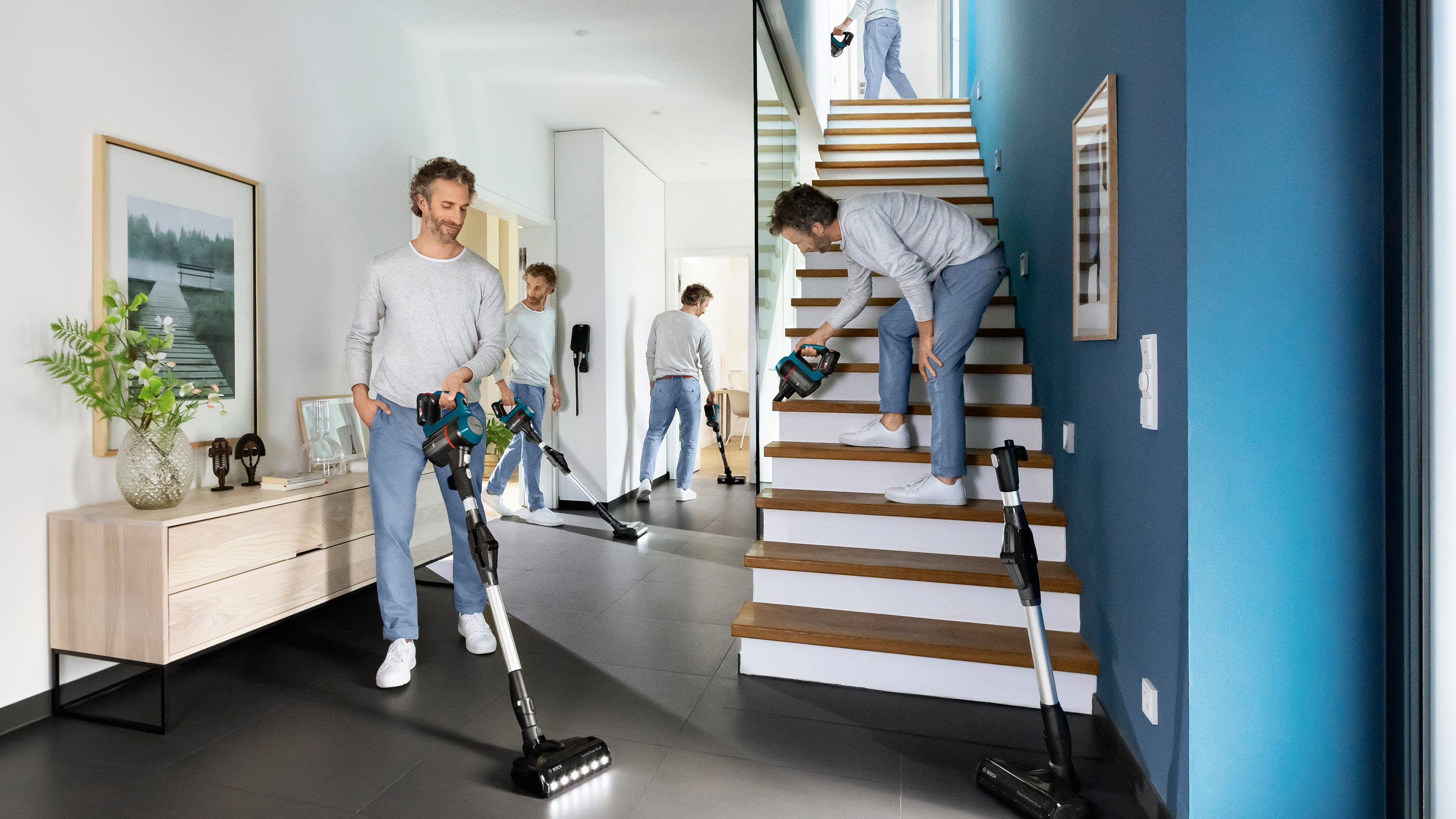 Mann saugt das Haus. Er saugt Treppen und Boden mit einem kabellosen Staubsauger. Er trägt einen grauen Pullover und blaue Hose.