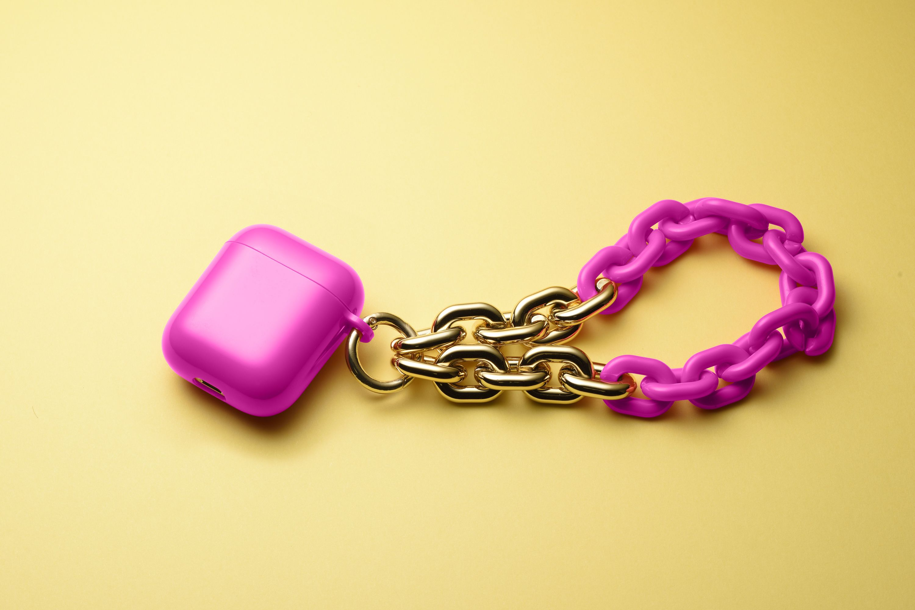 Étui AirPods rose attaché à une chaîne dorée et un bracelet chaîne rose, posé sur une surface jaune.