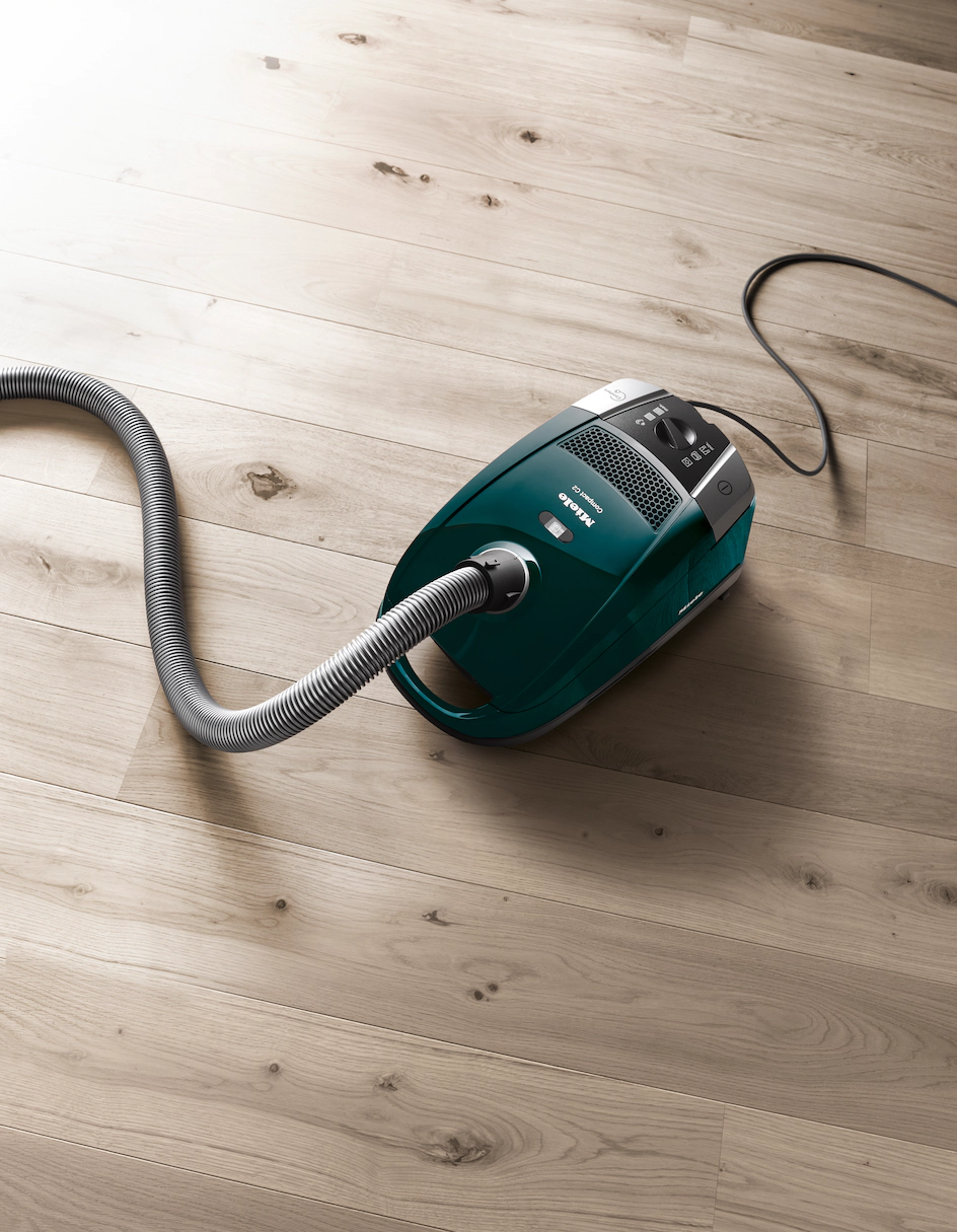 Een turquoise stofzuiger met een slang ligt op een lichte houten vloer.