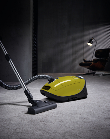 Un aspirateur jaune sur un tapis, avec une chaise et une lampe en arrière-plan.