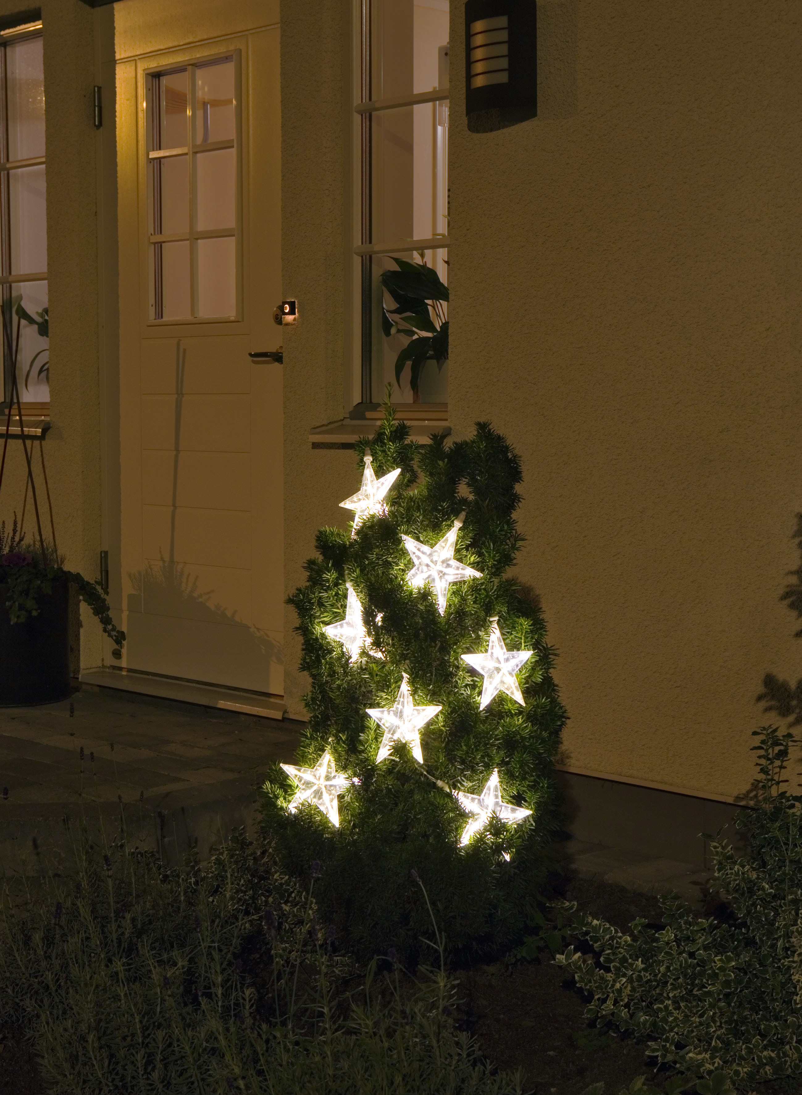 Ein kleiner Weihnachtsbaum aus Sternlichtern steht nachts vor einem Haus.