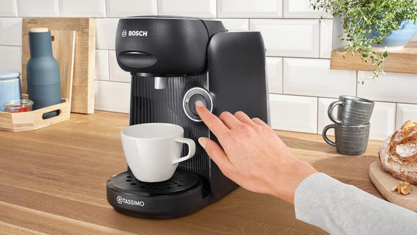 Eine schwarze Kaffeemaschine mit einer weißen Tasse, eine Hand drückt einen Knopf, Holzarbeitsplatte.