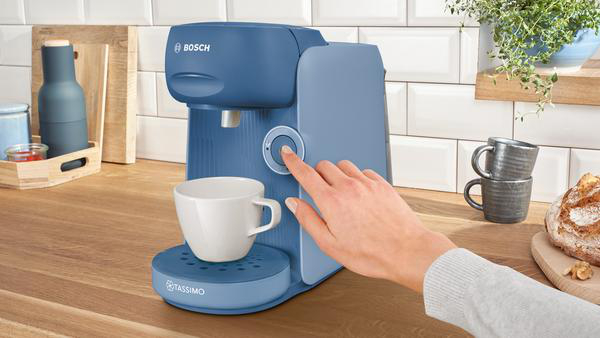 Blaue Kaffeemaschine mit einer Hand, die einen Knopf drückt, Tasse und Brot sichtbar.