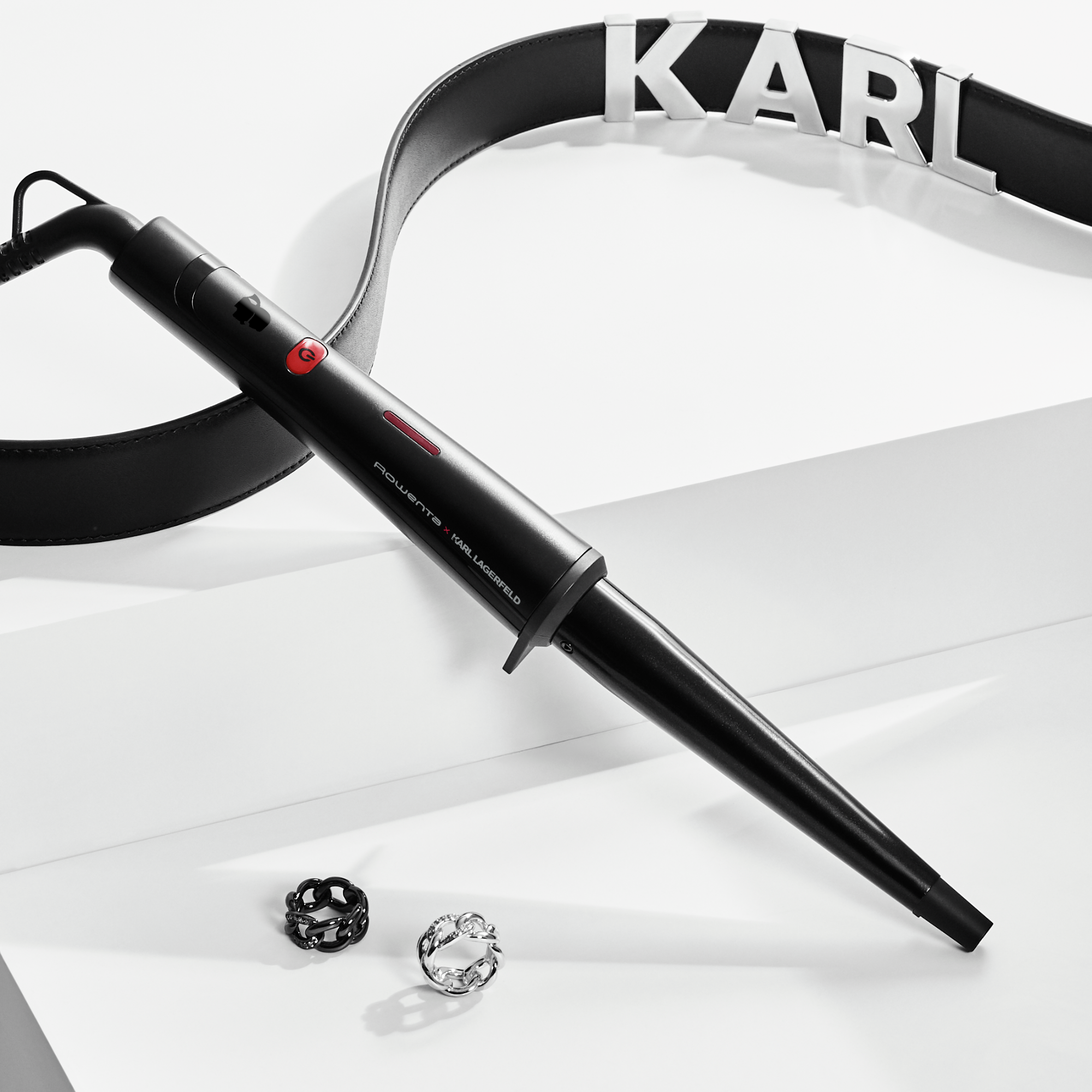 Rizador negro, dos anillos y un cinturón con letras KARL sobre superficie blanca.