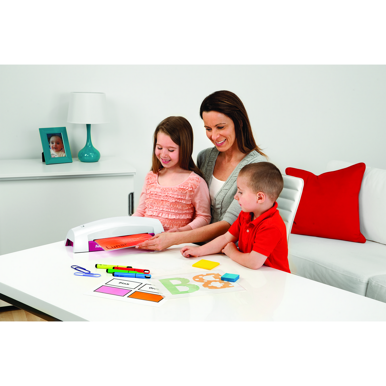 Una mujer y dos niños están creando. Usan una plastificadora y suministros coloridos en una mesa.