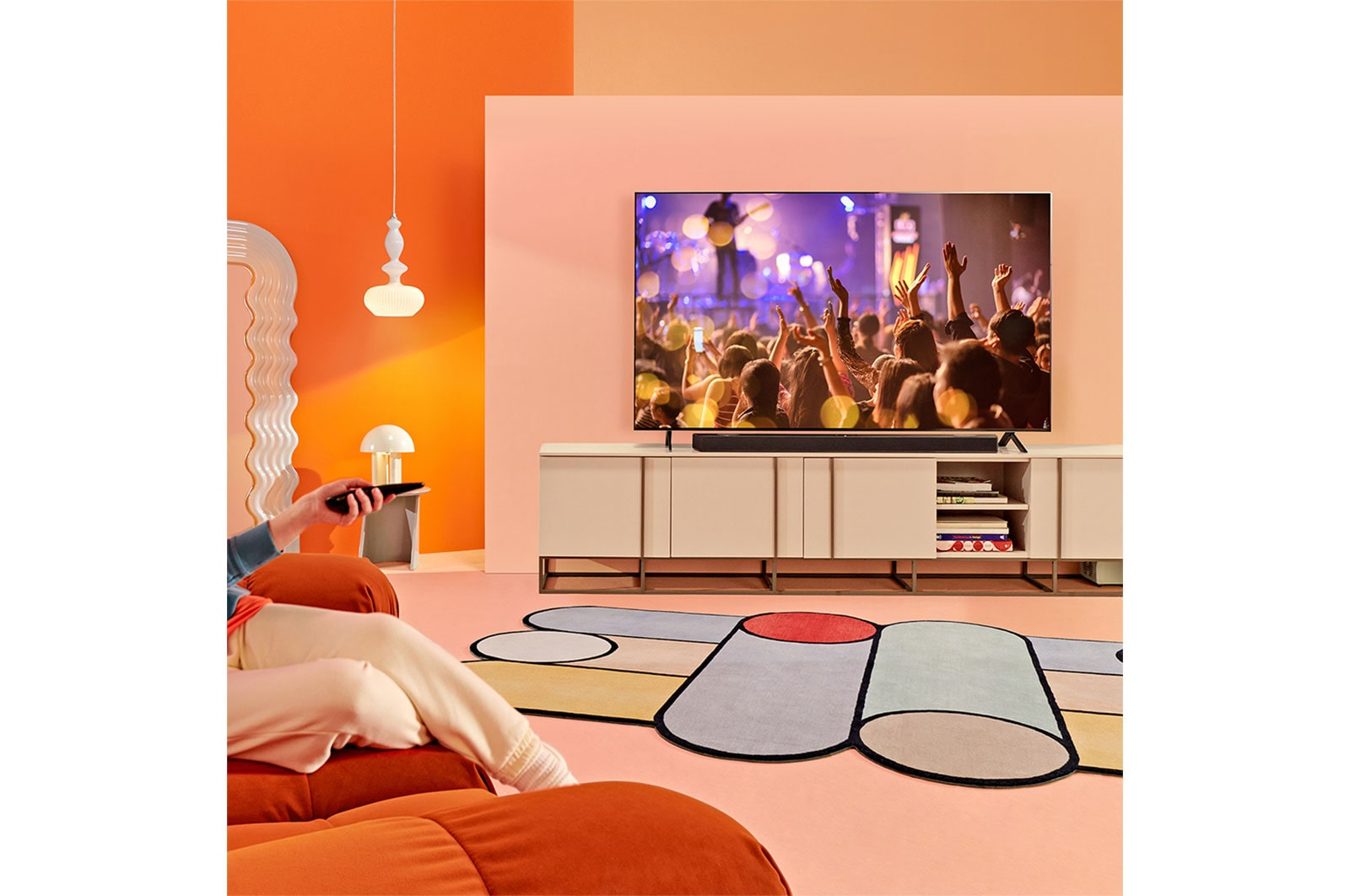 Oturma odası sahnesi. Konser gösteren TV, altında soundbar. Ön planda kanepe ve halı.