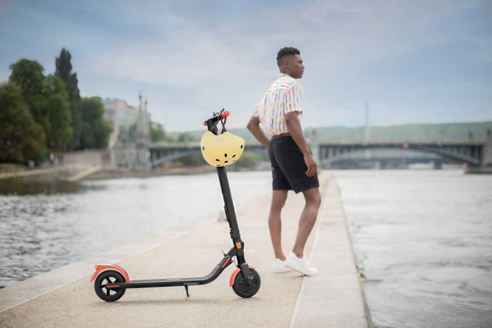 Bir adam, arka planda bir köprü ile, bir nehrin yanında bir scooter'ın yanında duruyor.
