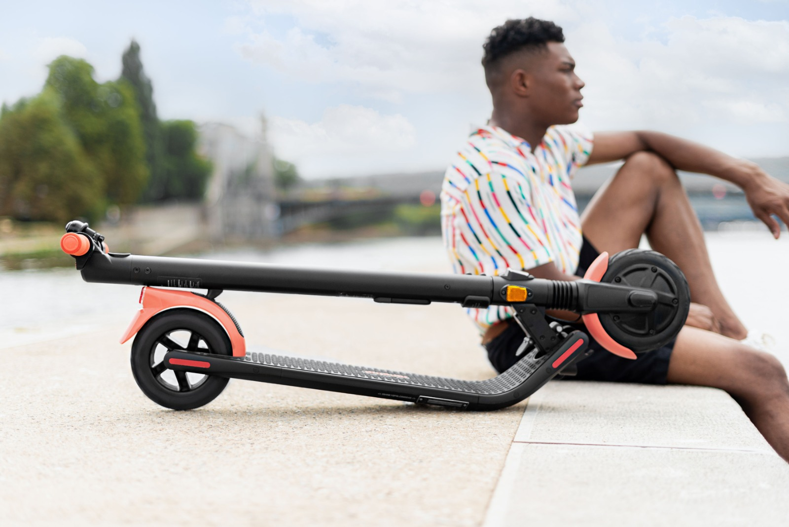 Bir adam, bulutlu bir gökyüzü ile suyun yanında katlanmış pembe ve siyah bir elektrikli scooter'ın yanında oturuyor.