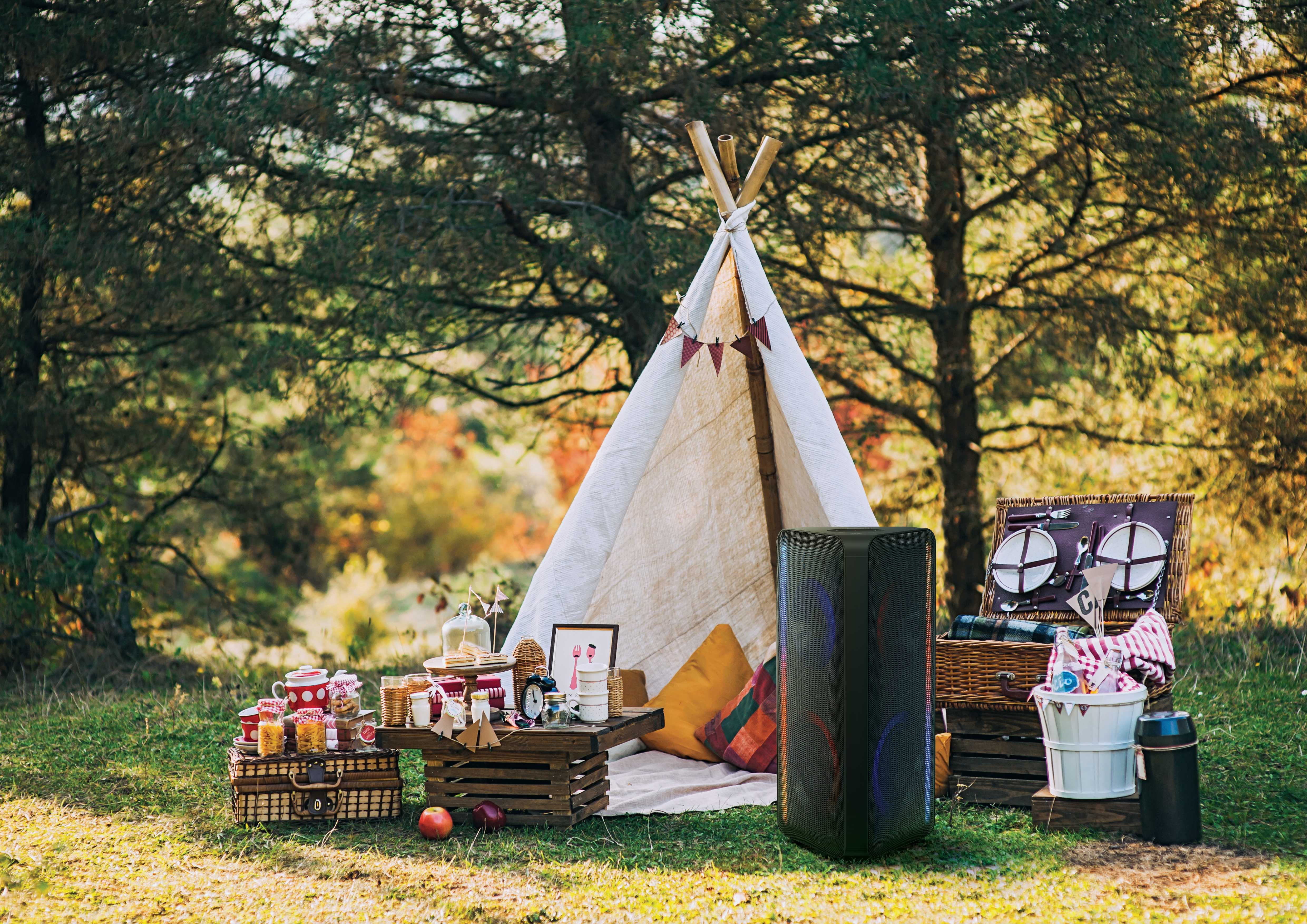 Picknick-Szene in der Natur. Zelt, Lautsprecher, Essen, Getränke und eine Decke auf Gras mit Bäumen im Hintergrund.