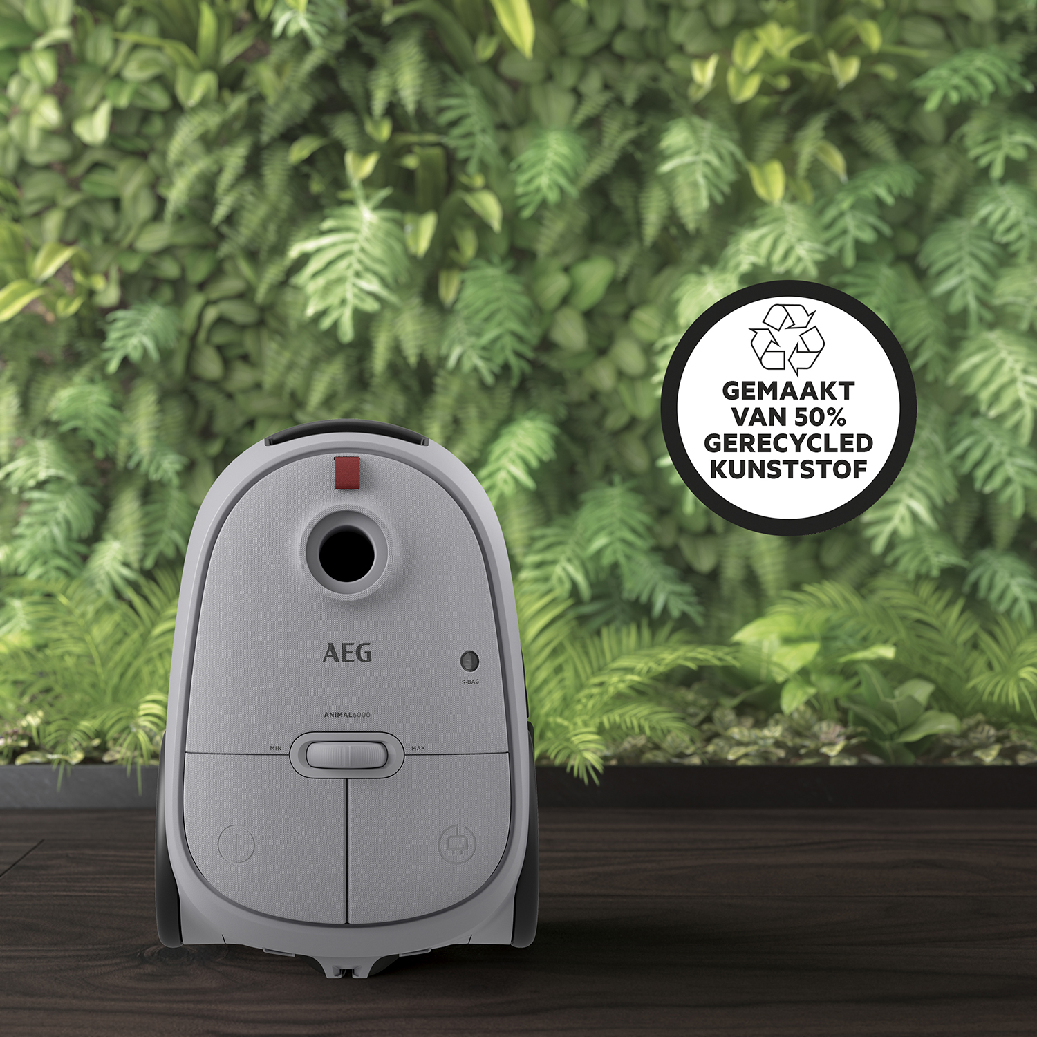 A gray vacuum cleaner stands in front of a wall of green plants. A label is visible.