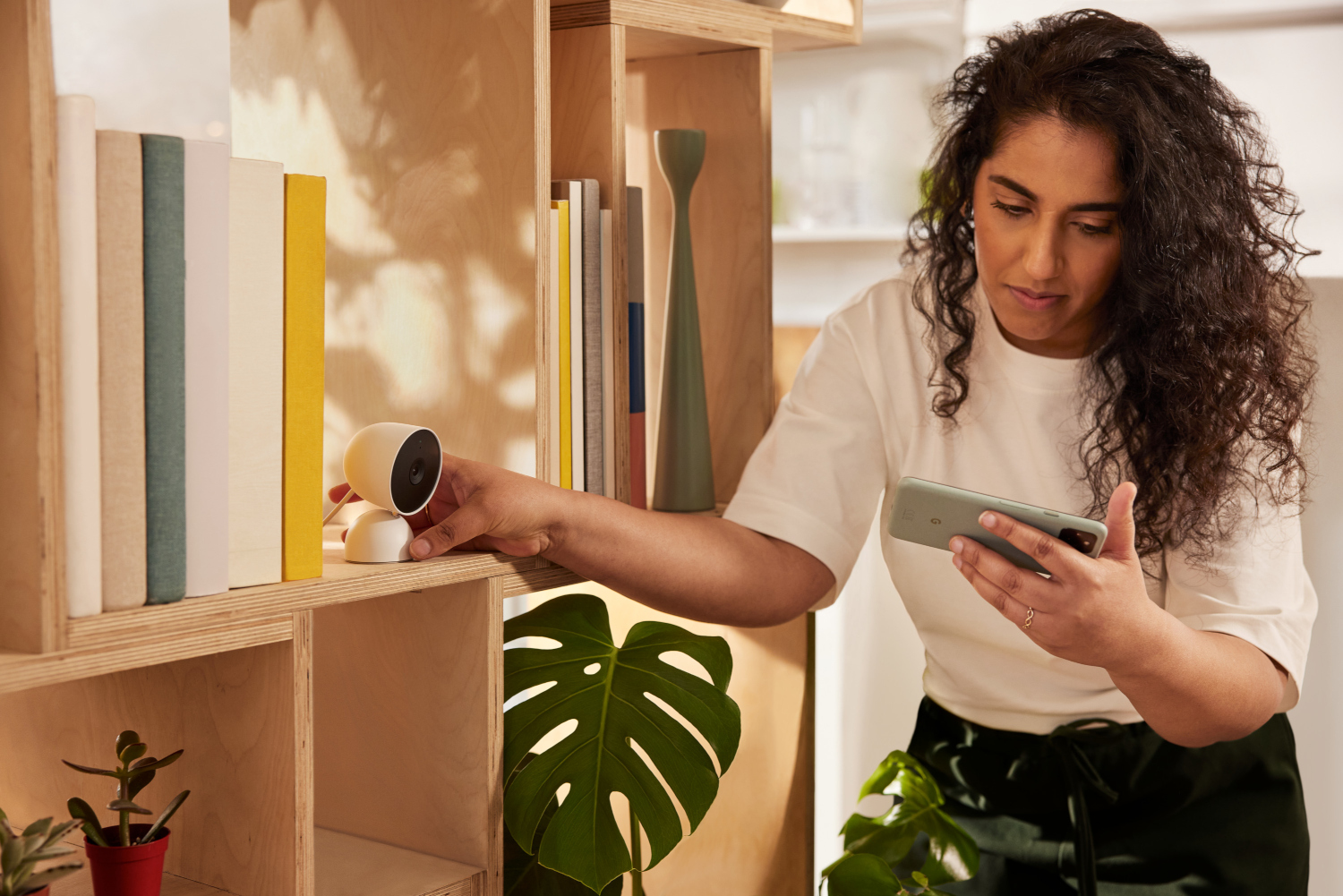 Une femme installe une caméra de sécurité sur une étagère avec des livres et une plante. Tenant un téléphone.